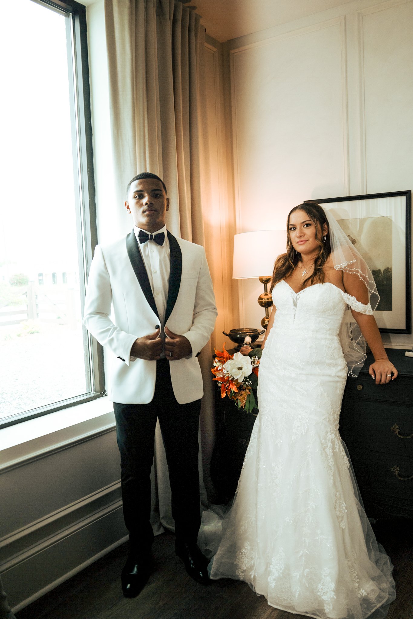 A groom in a white tuxedo with a black bow tie standing next to a bride in a white wedding dress indoors near a window, a lamp, and a small black dresser with a floral bouquet on top.