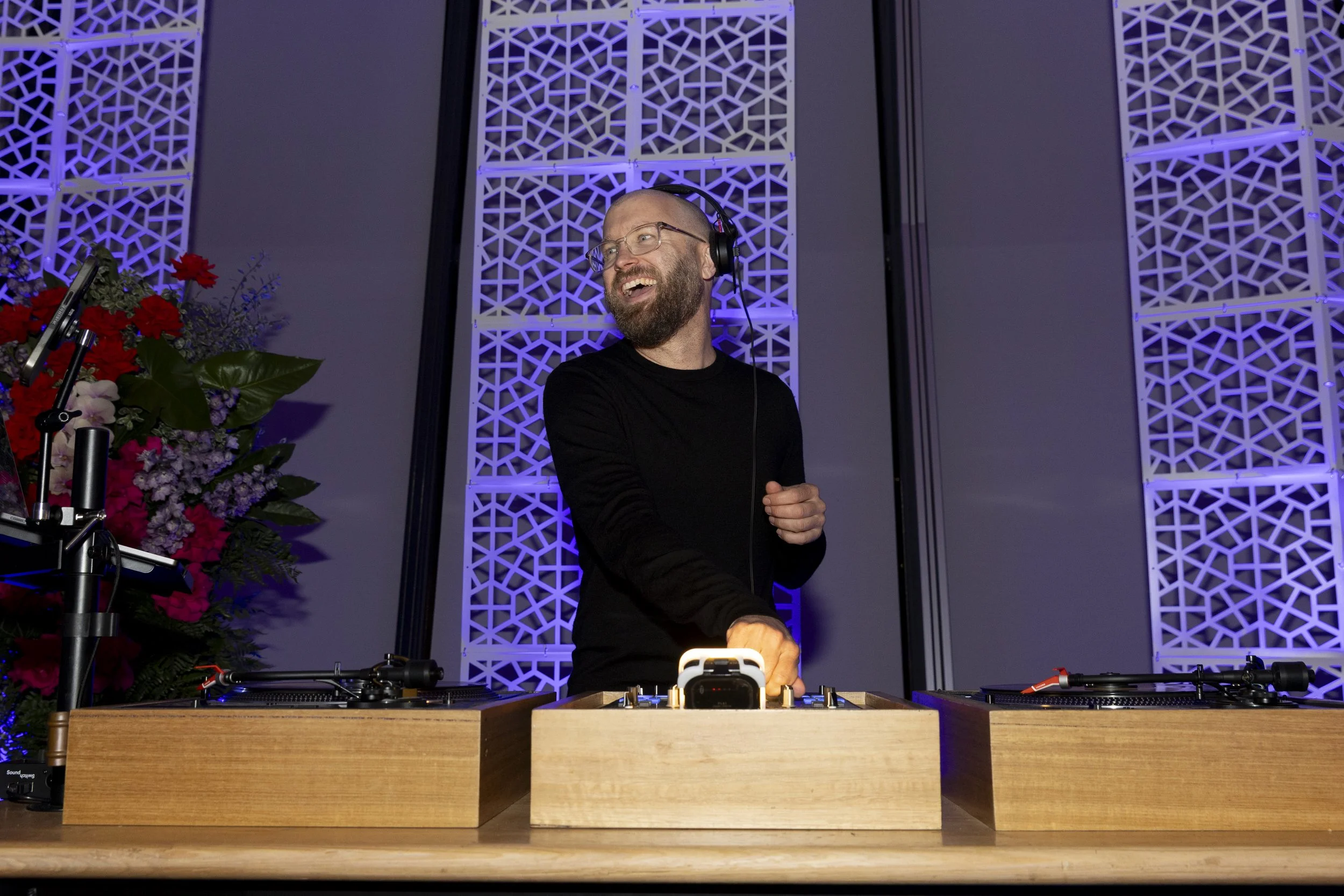 Bald dj wearing headphones and standing behind a wooden desk with vinyl turntables
