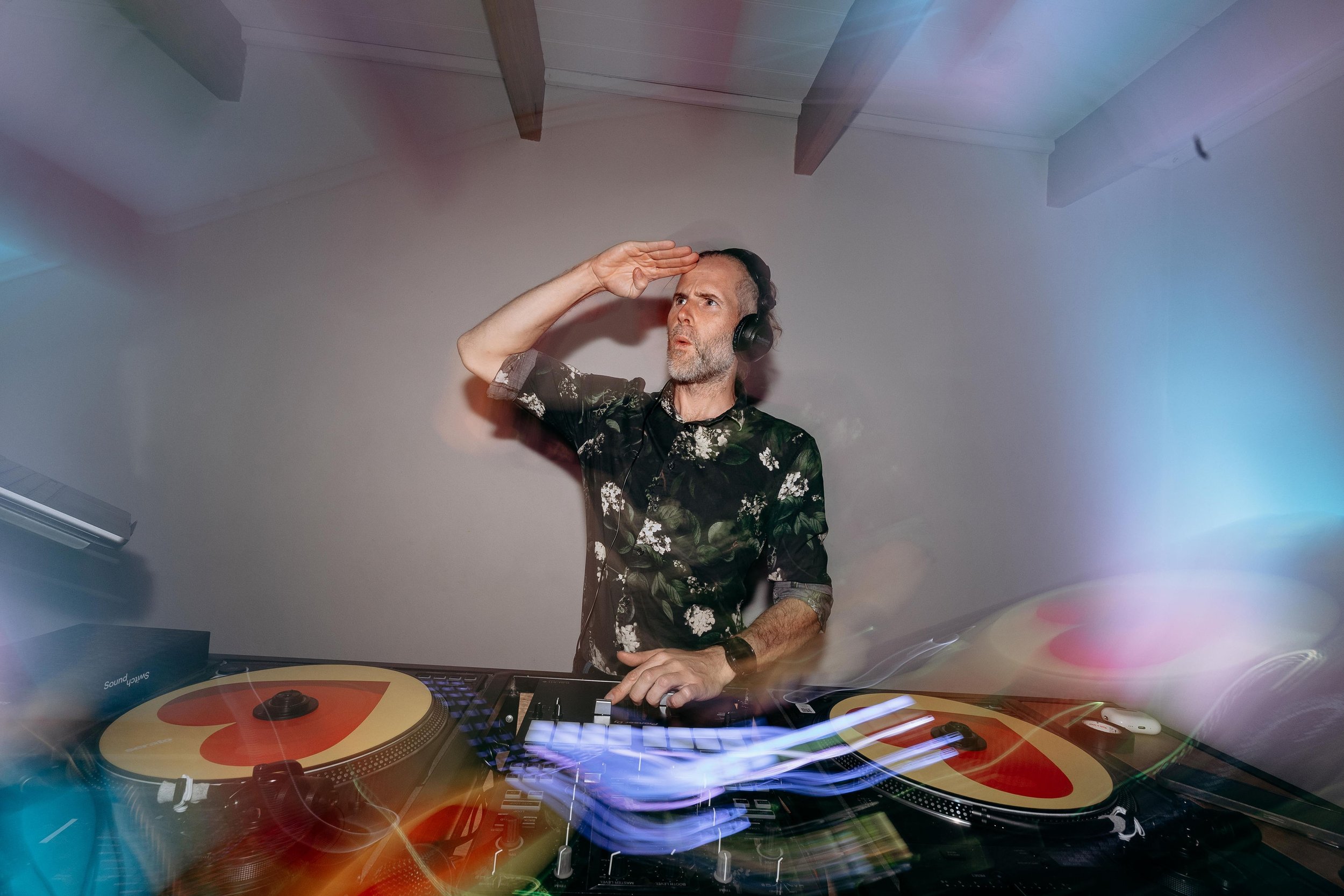 a dj with long hair grinning whilst standing behind vinyl turntables