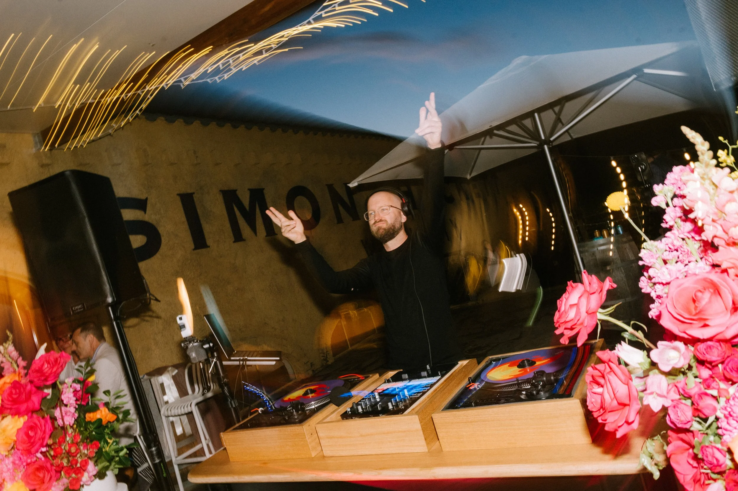 A male dj wearing glasses standing behind vinyl turntables and pointing in the air with both hands