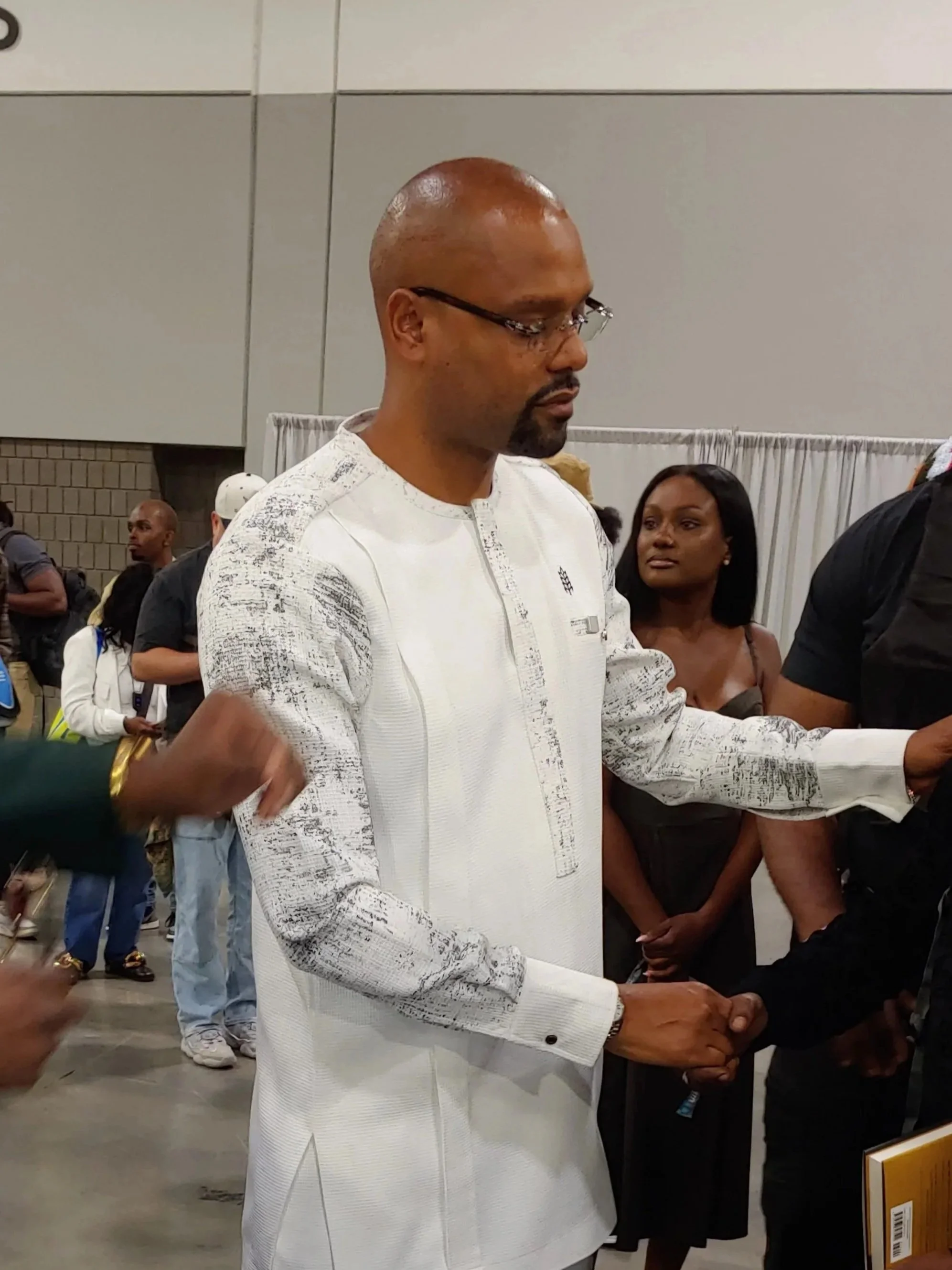 Rashad Bilal in a white traditional African outfit with black and silver patterns, wearing glasses, shaking hands with another person at the Invest Fest EYL book signing.