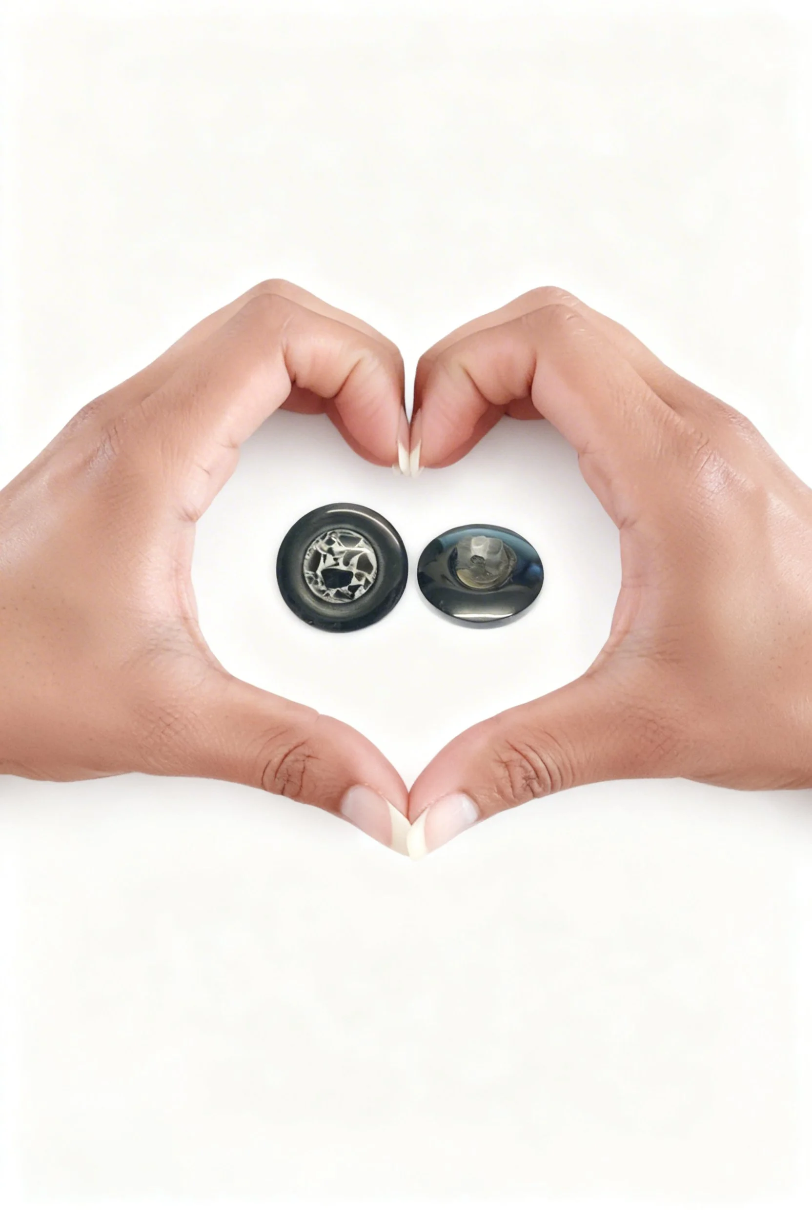 Two hands coming together in the shape of a heart that encloses 2 vintage black buttons one top facing and the other bottom facing to show the rear see through shank of the button.