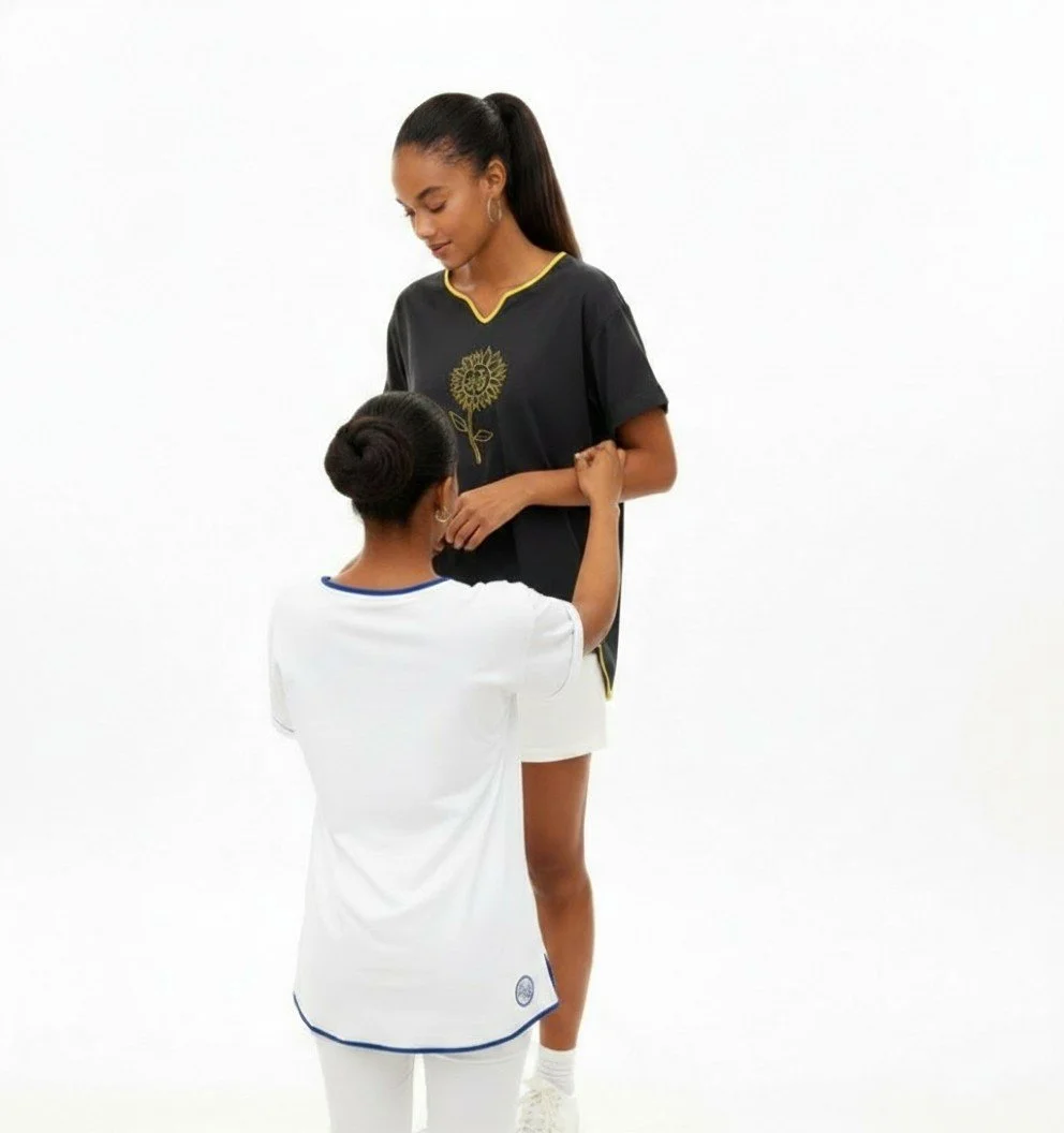 Two models standing together in a photoshoot. One model is on a platform looking down while the other model reaches up to make an adjustment to the t-shirt she is wearing.