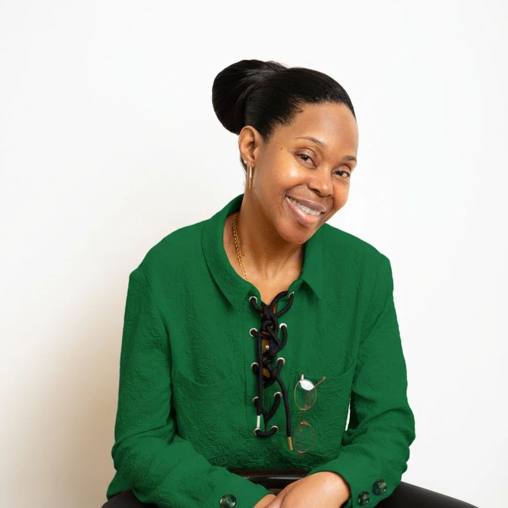 A woman smiling, wearing a green jacquard blouse, with glasses hanging from the patch pocket of her shirt, leaning forward against a bar stool.