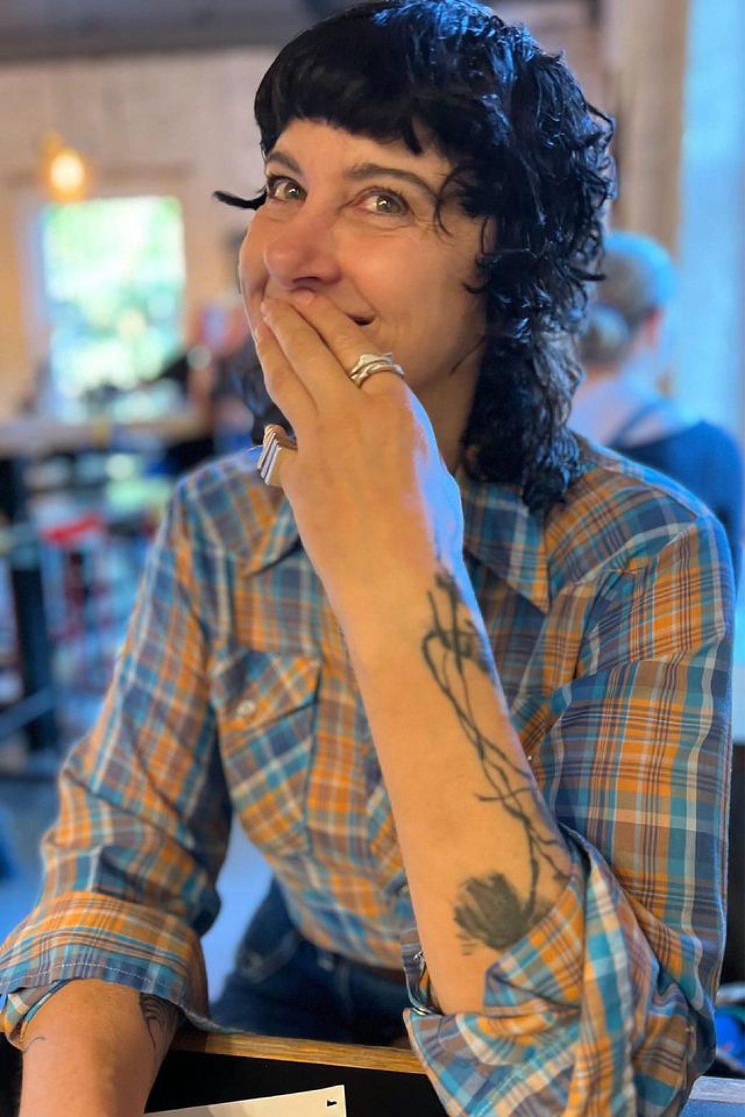 Jane Durlacher, Graphic Designer, profile picture with short black hair wearing a plaid shirt, hand covering mouth, showing a tattoo on forearm, smiling indoors.