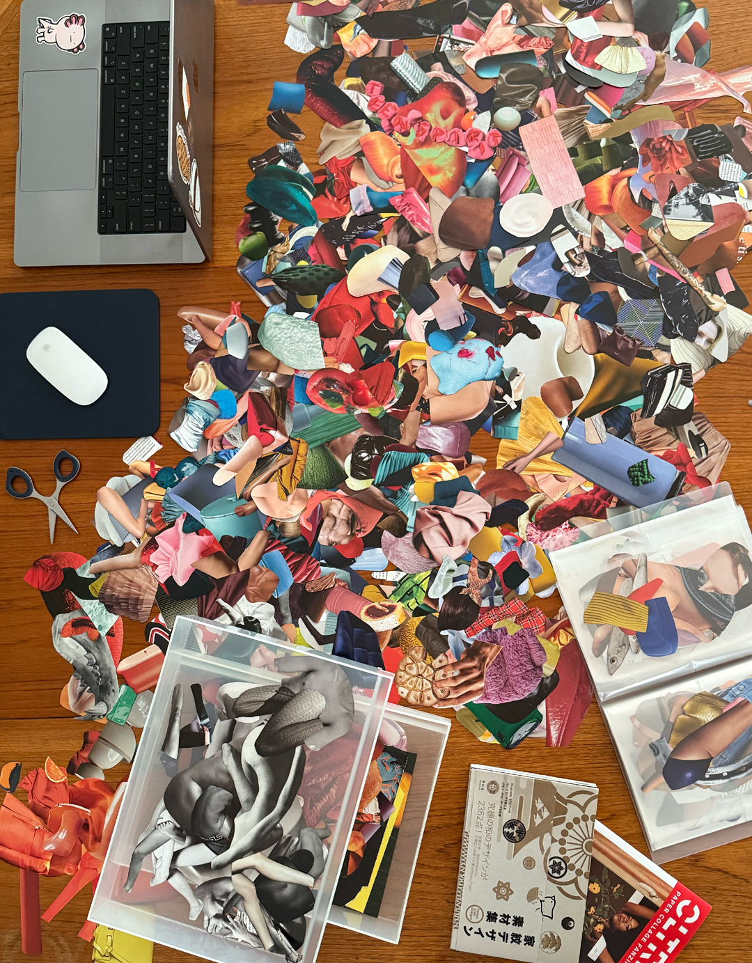 A wooden table cluttered with a laptop, a computer mouse on a pad, scissors, and a large assortment of magazine cutouts ready to be collaged. The cutouts feature various images and are spread haphazardly. There is also a Japanese design book in view.