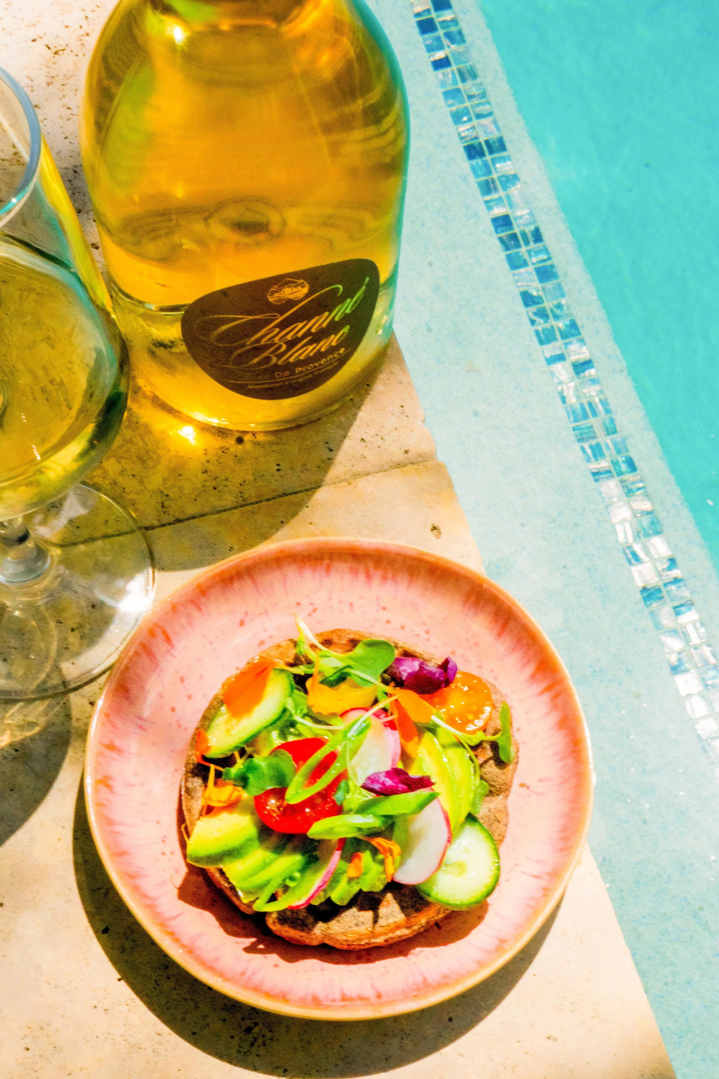 A plate with avocado toast topped with mixed vegetables and greens, beside a pool with two glasses of white wine.