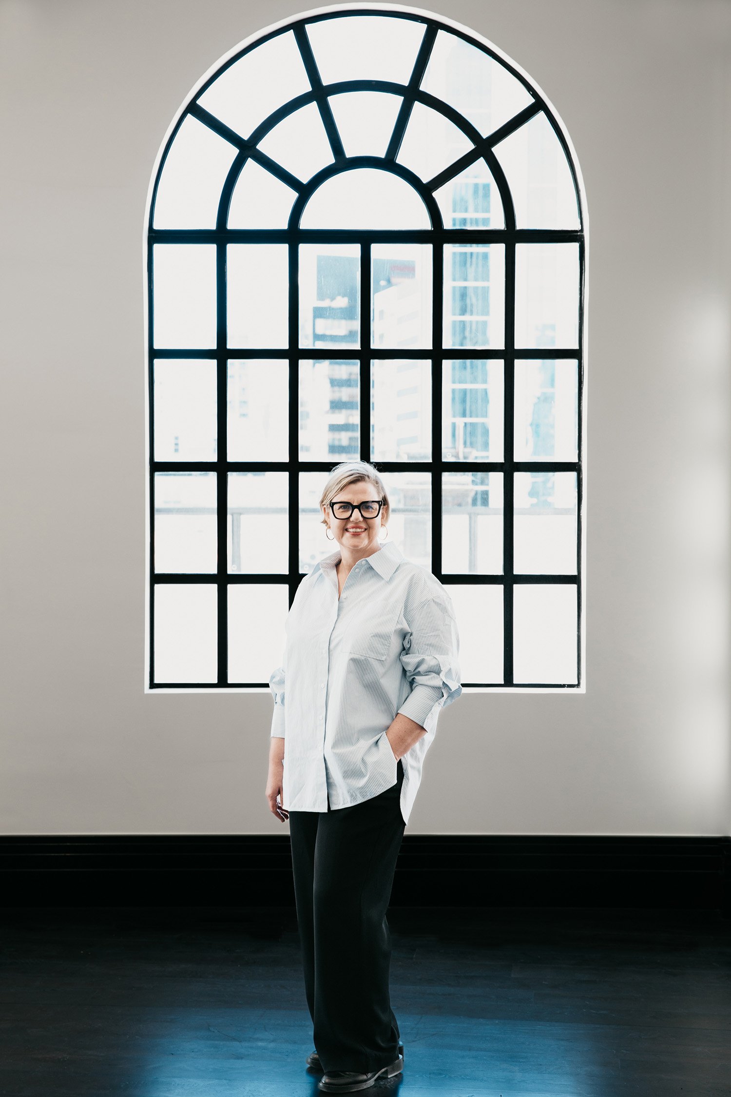 A woman with short blonde hair and glasses, wearing a white button-up shirt and black pants, stands in front of a large arched window with a cityscape view.