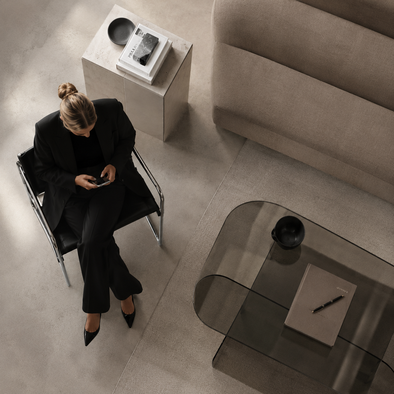 A woman in black business attire sitting on a black chair, looking at her phone in a modern, minimalist living room with a glass coffee table, a beige sofa, and a small side table with gray and black objects.