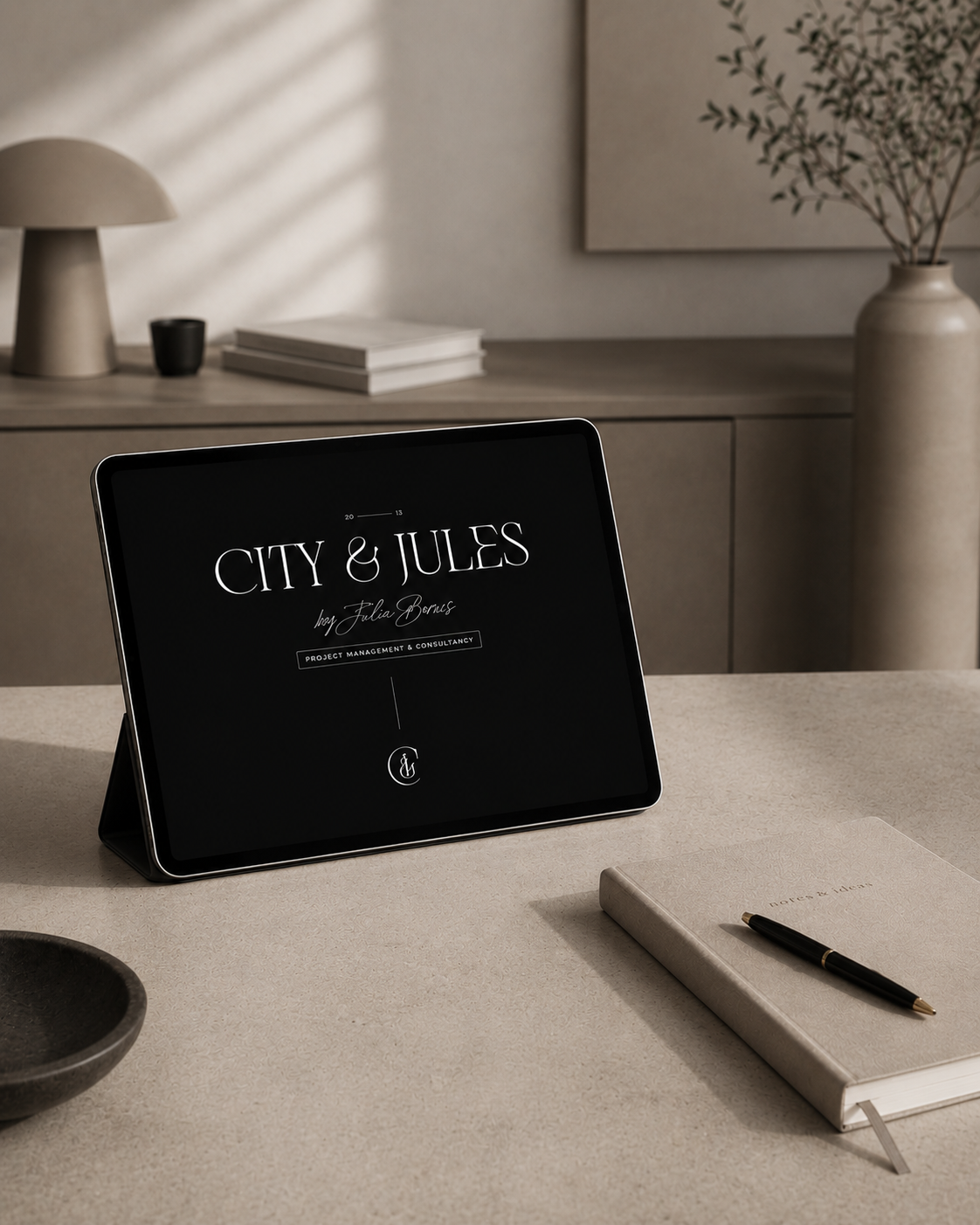A digital tablet on a desk displaying a project titled "City & Jules" by Julia Bonus. The desk has a notebook with a pen, a dark bowl, and is set in a neutral-toned room with minimalist decor including a lamp, books, and a vase with a leafy plant.