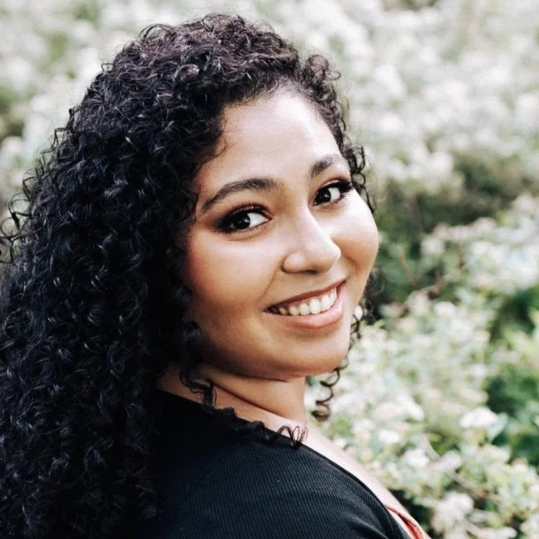 Headshot of Onella Piyatilake with long curly hair smiling among white flowering bushes.