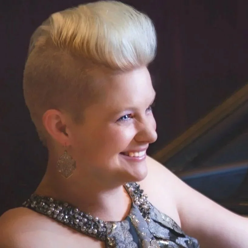 Headshot of Danielle Marie Steele smiling with short platinum blonde hair, wearing a glamorous dress with embellishments, earrings, and playing a grand piano in a dimly lit room.