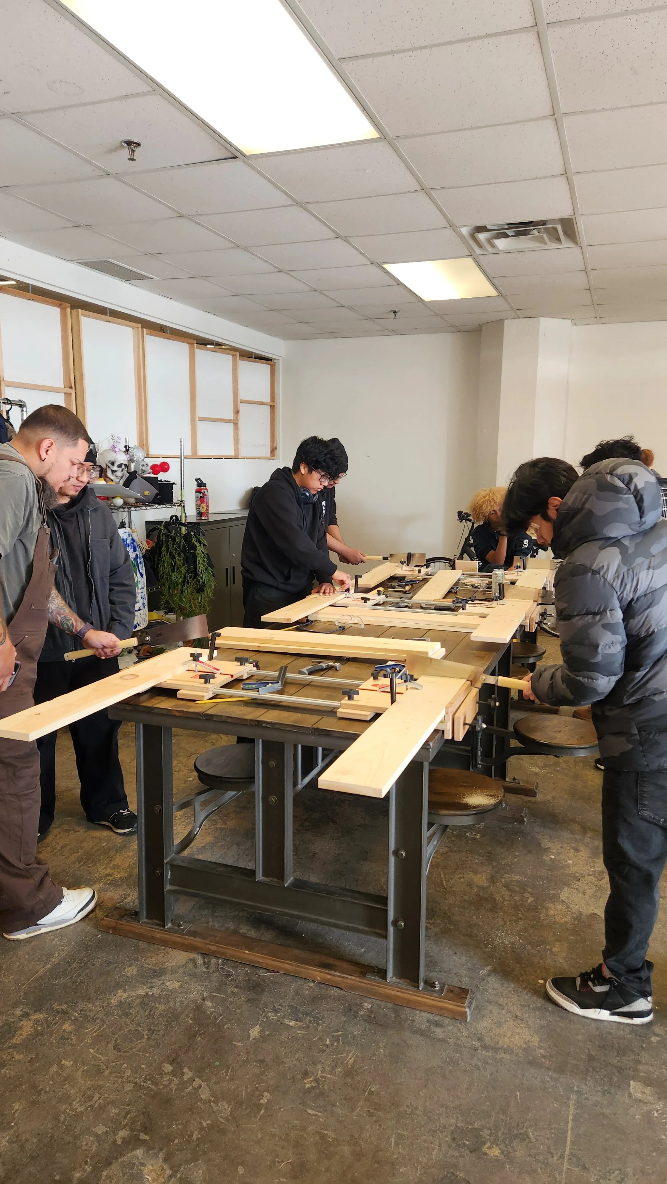 People working on woodworking projects at a workshop table with clamps and wood pieces.