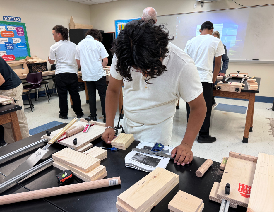 Classroom with students working on woodworking projects at tables, some cutting and assembling wooden pieces, with a whiteboard and educational posters in the background.