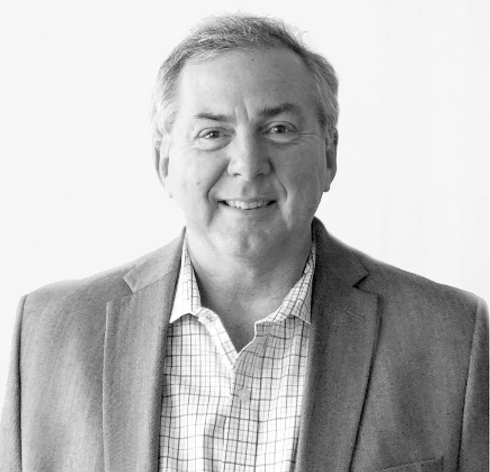 A black and white photo of a middle-aged man wearing a suit jacket and checkered shirt, smiling at the camera.