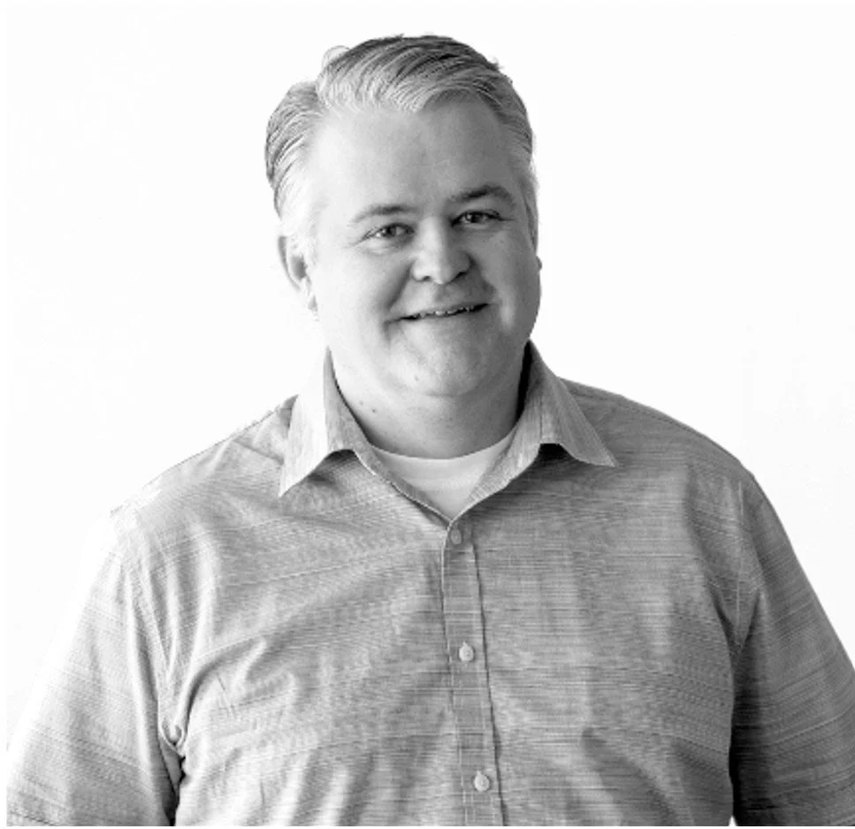 A black-and-white portrait of a smiling man with light-colored hair wearing a button-up shirt.