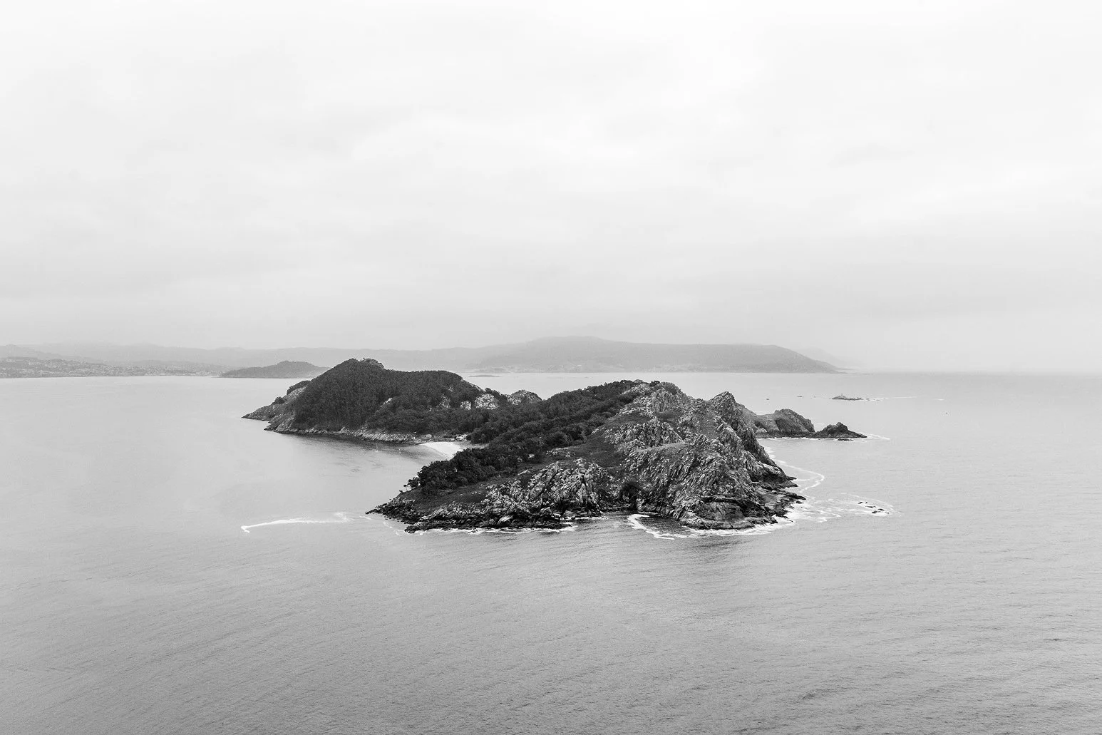 Black and white photo of a small island with rocky shores and dense trees in the ocean, with a distant landmass on the horizon under cloudy sky.