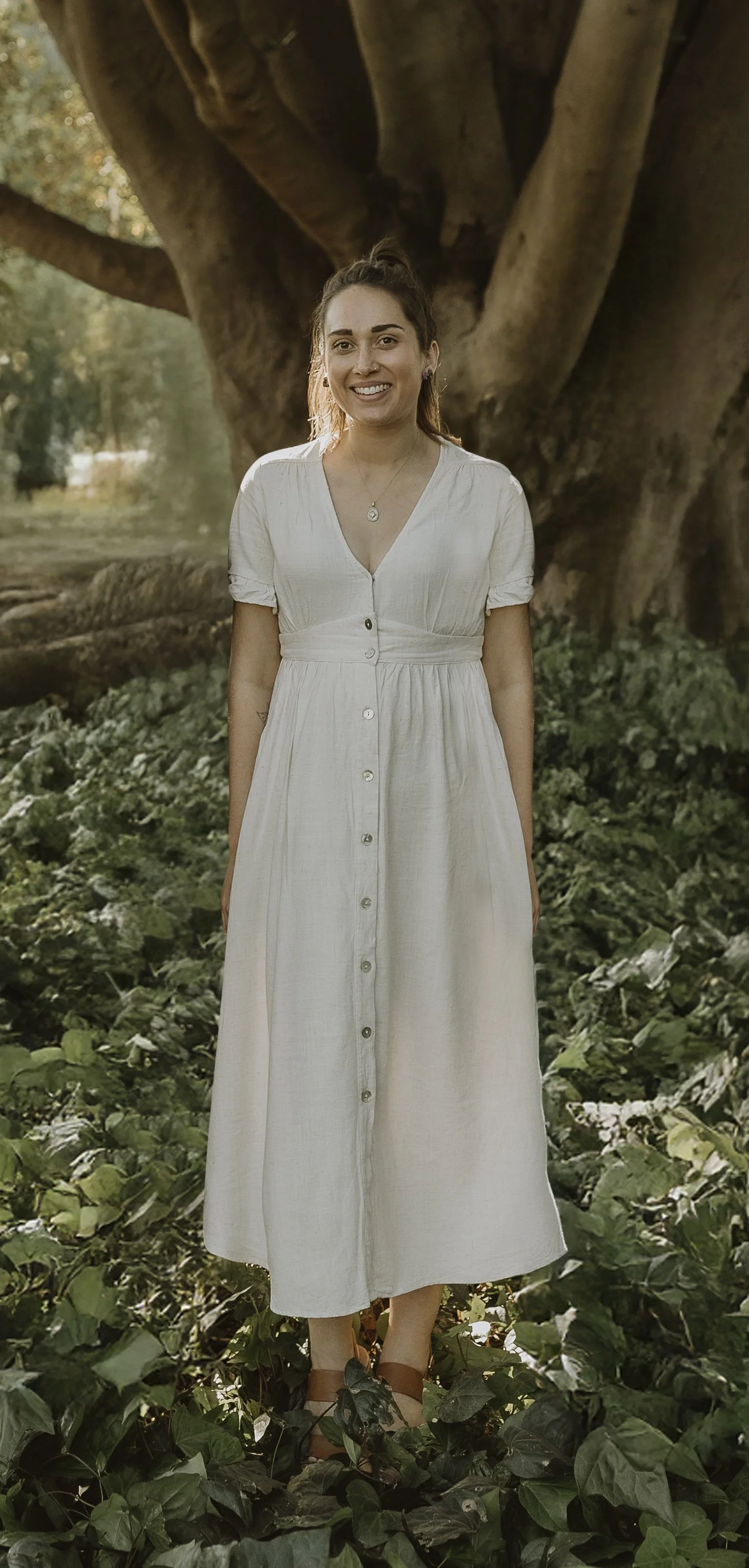 A woman in a white dress standing outdoors among green foliage with a large tree in the background.