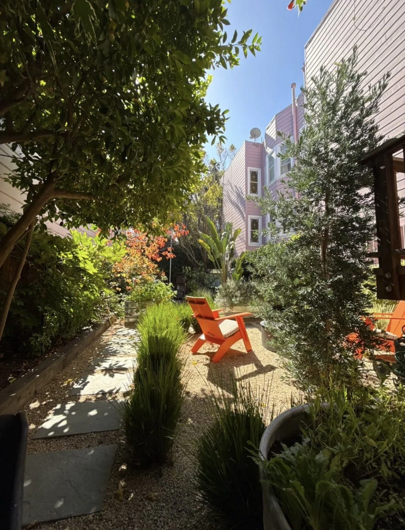 A cozy outdoor garden with lush greenery and trees, orange wooden chairs, a stone pathway, and a multi-story pink building in the background on a sunny day.