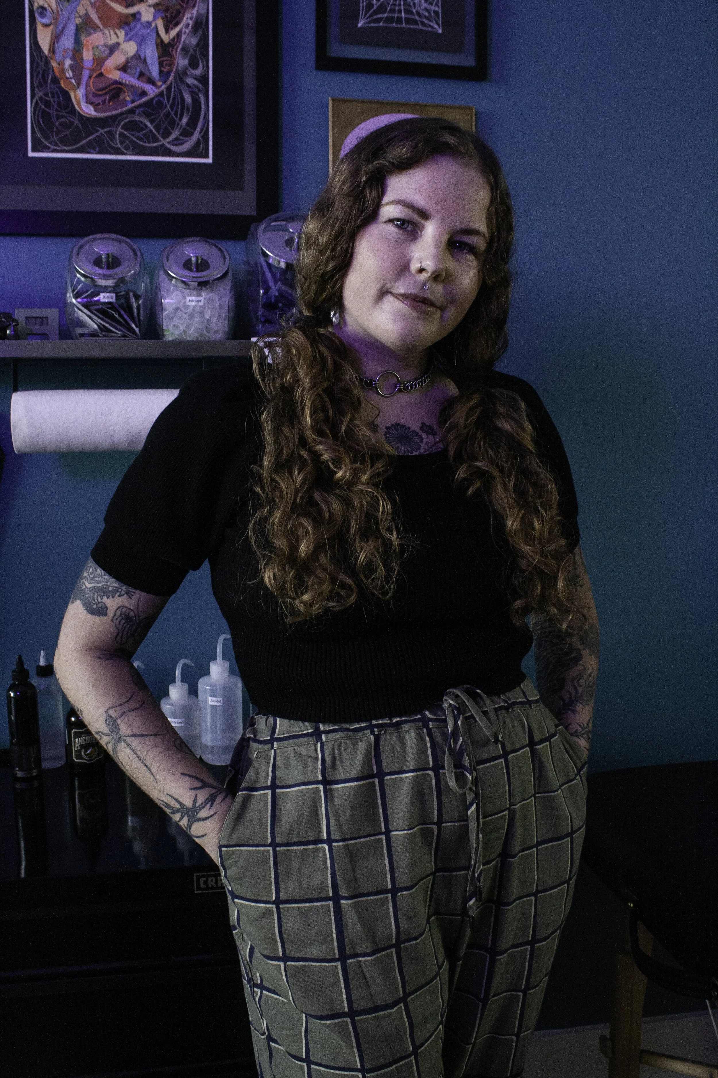 A young woman with long curly hair and tattoos on her arms stands in a tattoo parlor, smiling at the camera. She wears a black top, checkered pants, a choker necklace, and has a septum piercing. The background features tattoo supplies and colorful artwork on the walls.