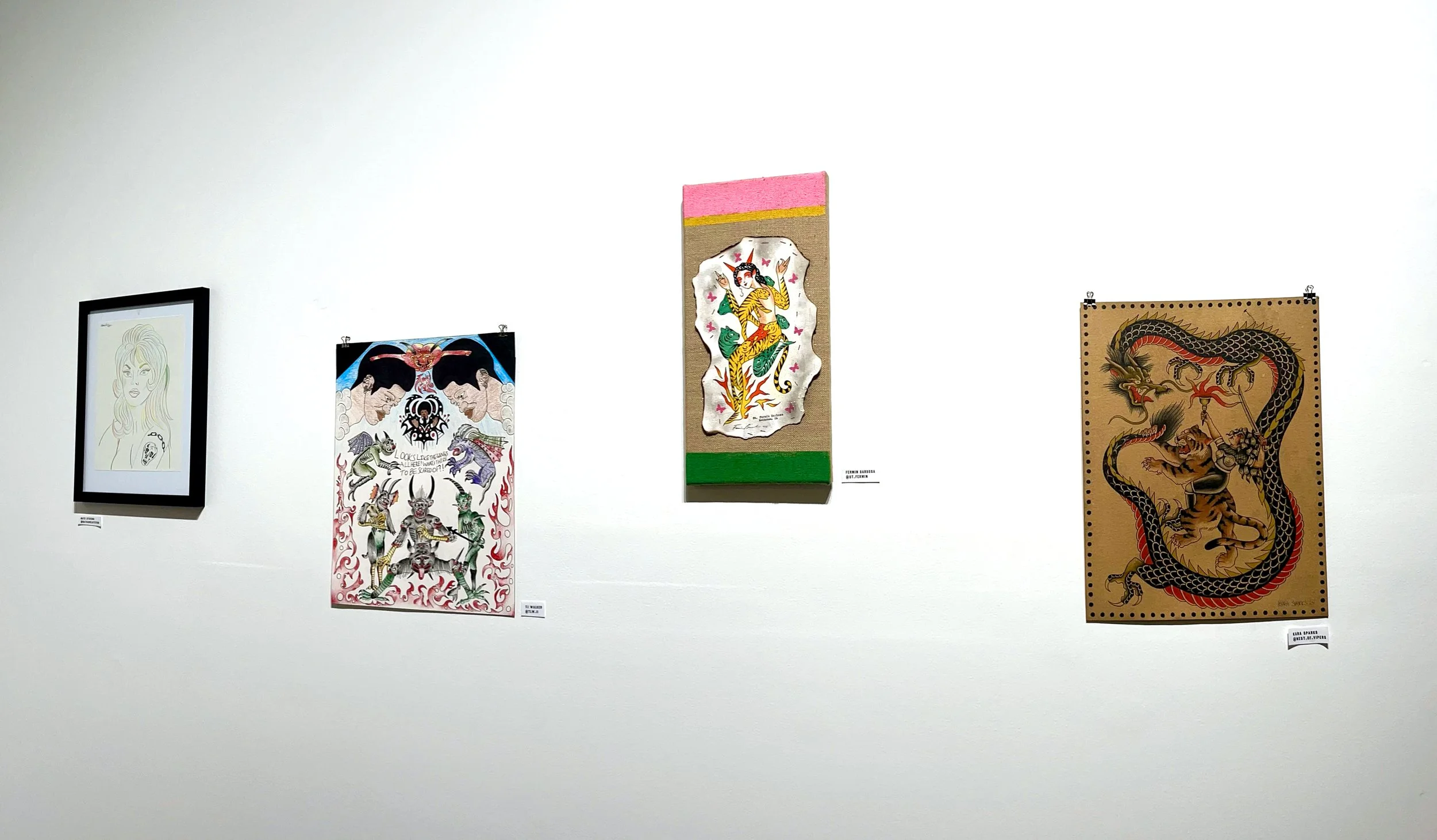 Art exhibition featuring five artworks: a minimalist line drawing of a woman, a colorful tattoo-style piece with mythical creatures and Japanese characters, a textile art of a woman in traditional clothing, a depicting a mythological creature, and a tattoo-inspired illustration with a dragon and tiger.