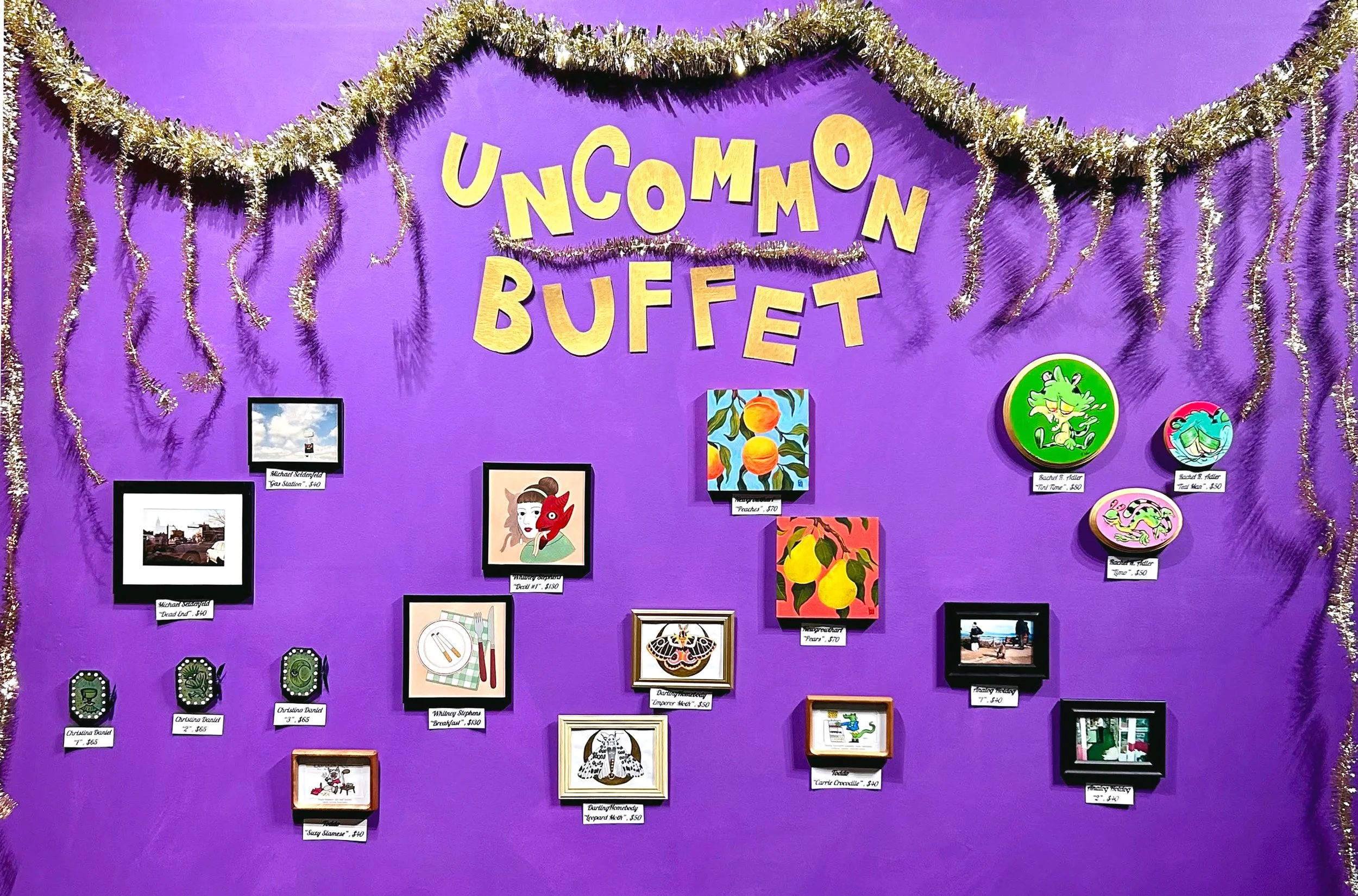 Purple wall decorated with silver tinsel and gold letter banners reading 'Uncommon Buffet'. Various framed pictures, illustrations, and ceramic items with price tags are displayed on the wall, showcasing artwork and handcrafted objects.