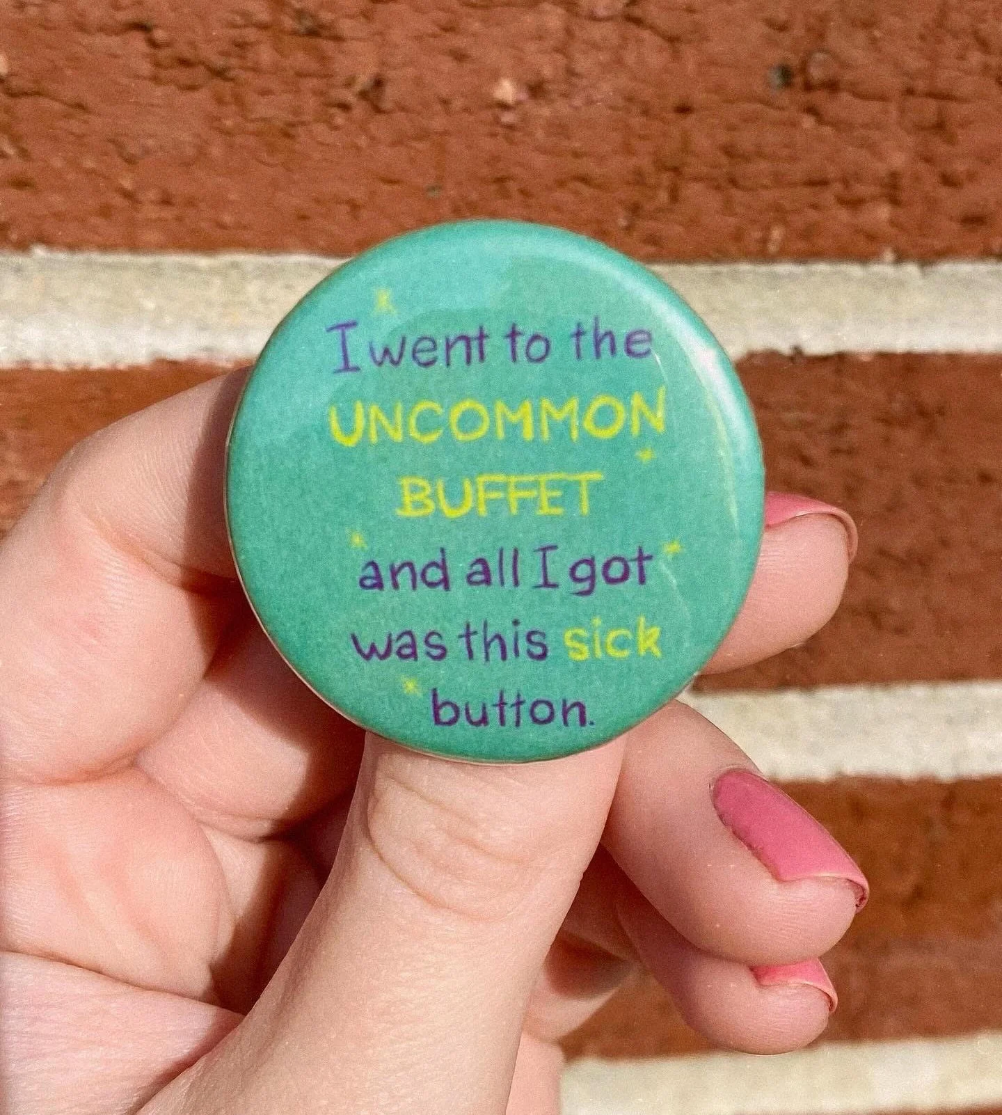 A round badge with the words 'I went to the UNCOMMON BUFFET and all I got was this sick button' written in colorful text against a green background with a brick wall behind it.