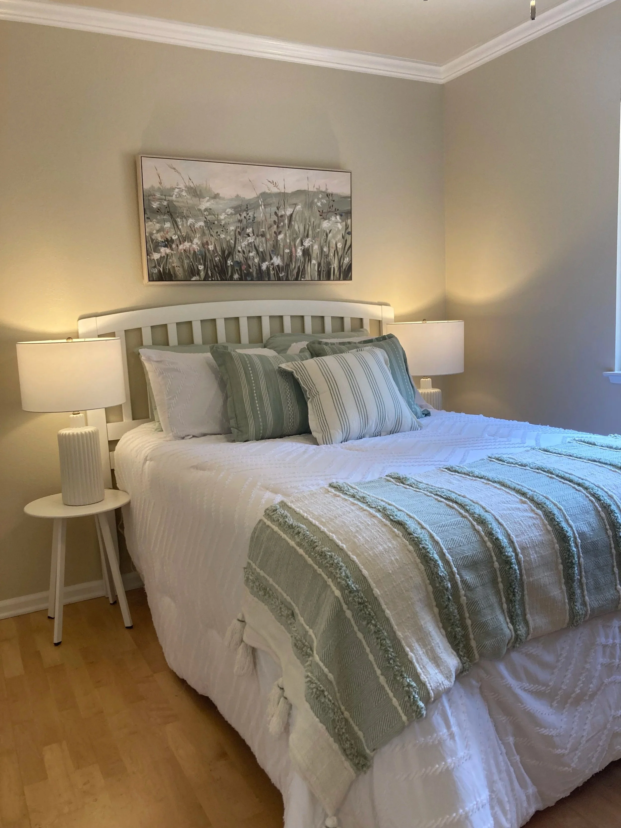 A neatly made bed with a white tufted comforter, multiple green pillows and a woven throw blanket at the foot, next to a window with white curtains and a lamp on a nightstand, in a cozy Foresthill California  bedroom.