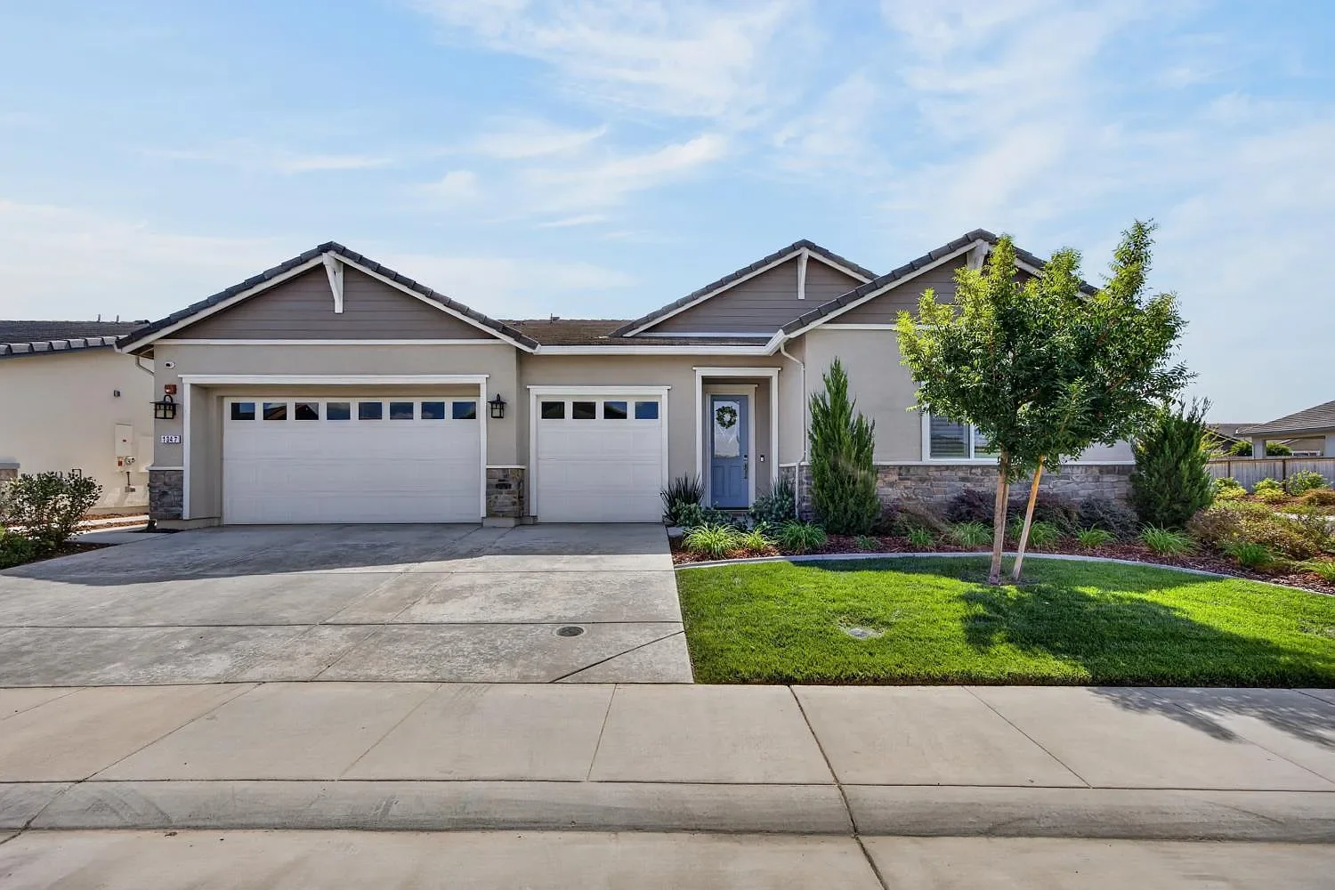 A modern single-story house with a two-car garage, front lawn, and landscaped garden with trees and shrubs, under a blue sky.