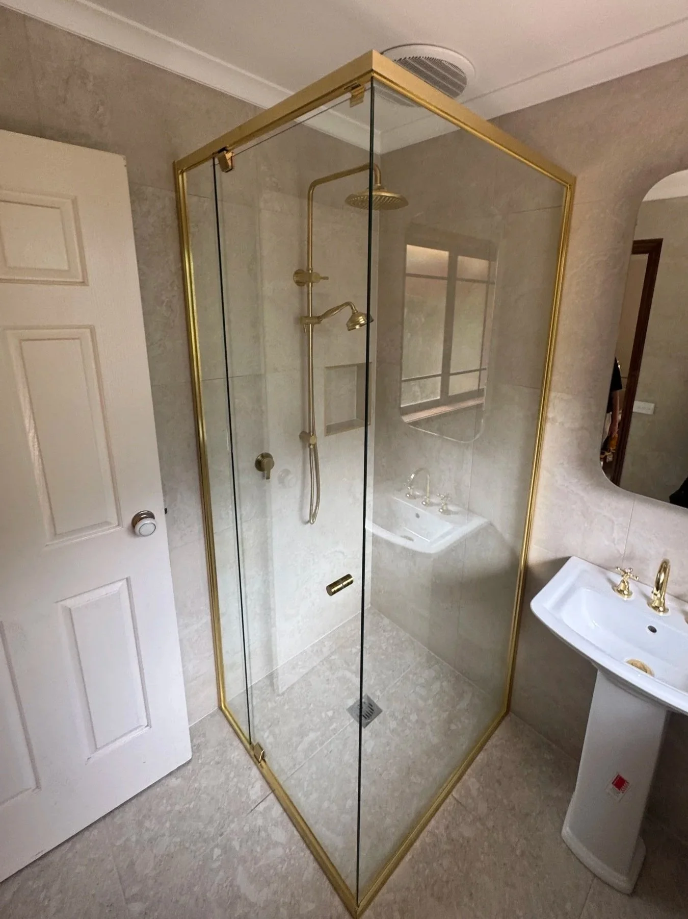 A bathroom shower with clear glass walls and gold framing, a gold showerhead and fixtures, a white pedestal sink, and a round mirror on the wall.