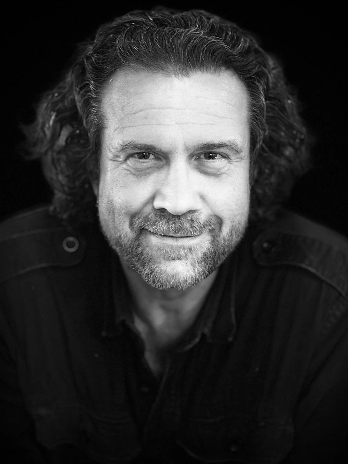 Black and white portrait of Stephan Doesinger with wavy, shoulder-length hair and a beard, smiling at the camera.