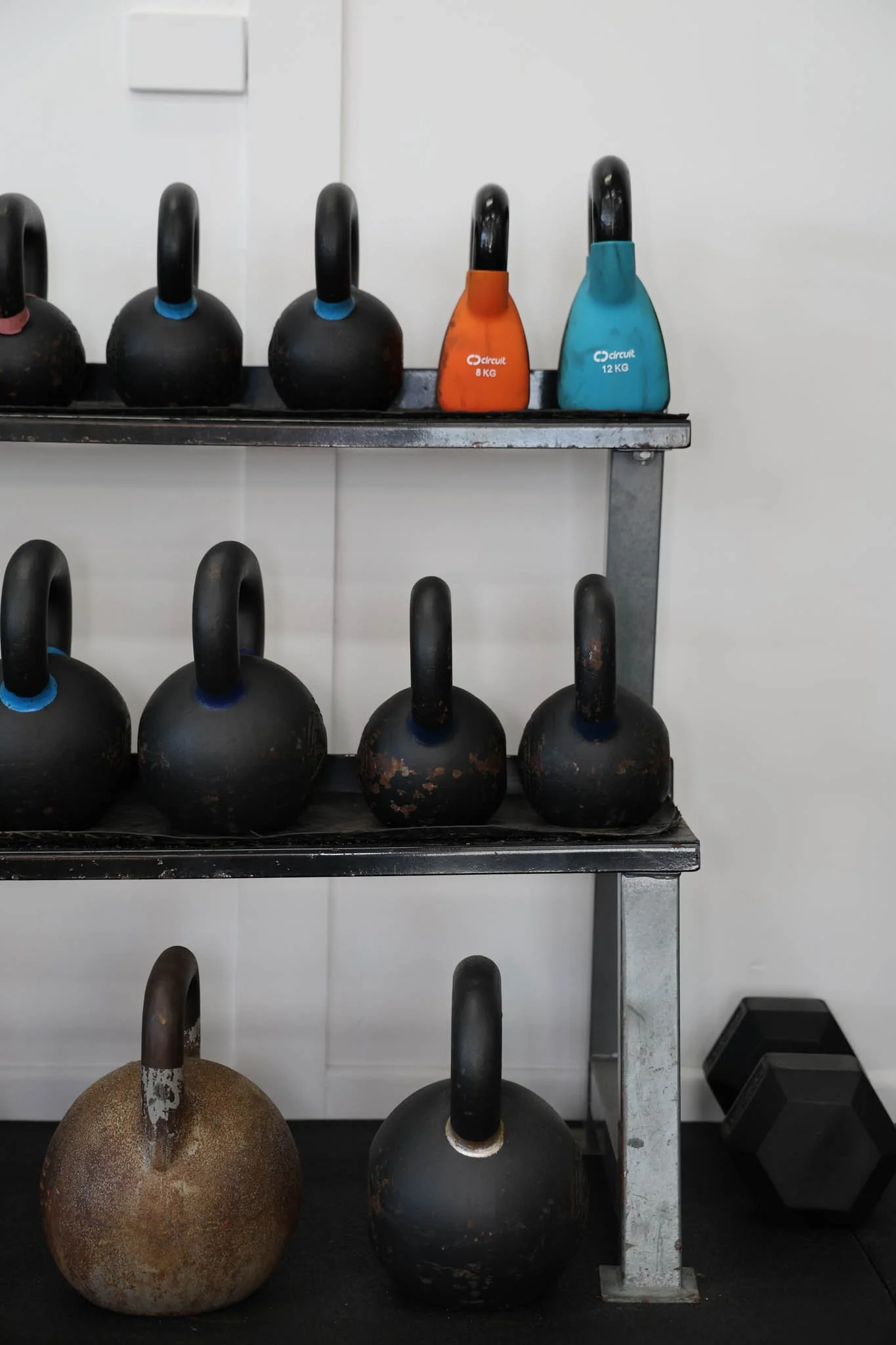 A metal rack holding multiple black kettlebells and two colorful kettlebells, one orange labeled 8 KG and one blue labeled 12 KG, against a white wall with electrical switch and black hex dumbbells on the floor.