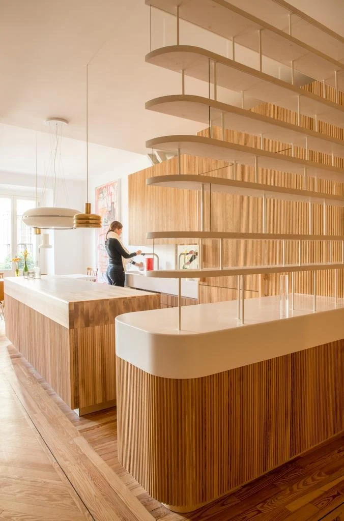 Modern kitchen with wooden cabinetry, a person standing at the sink, and curved, open shelving above.