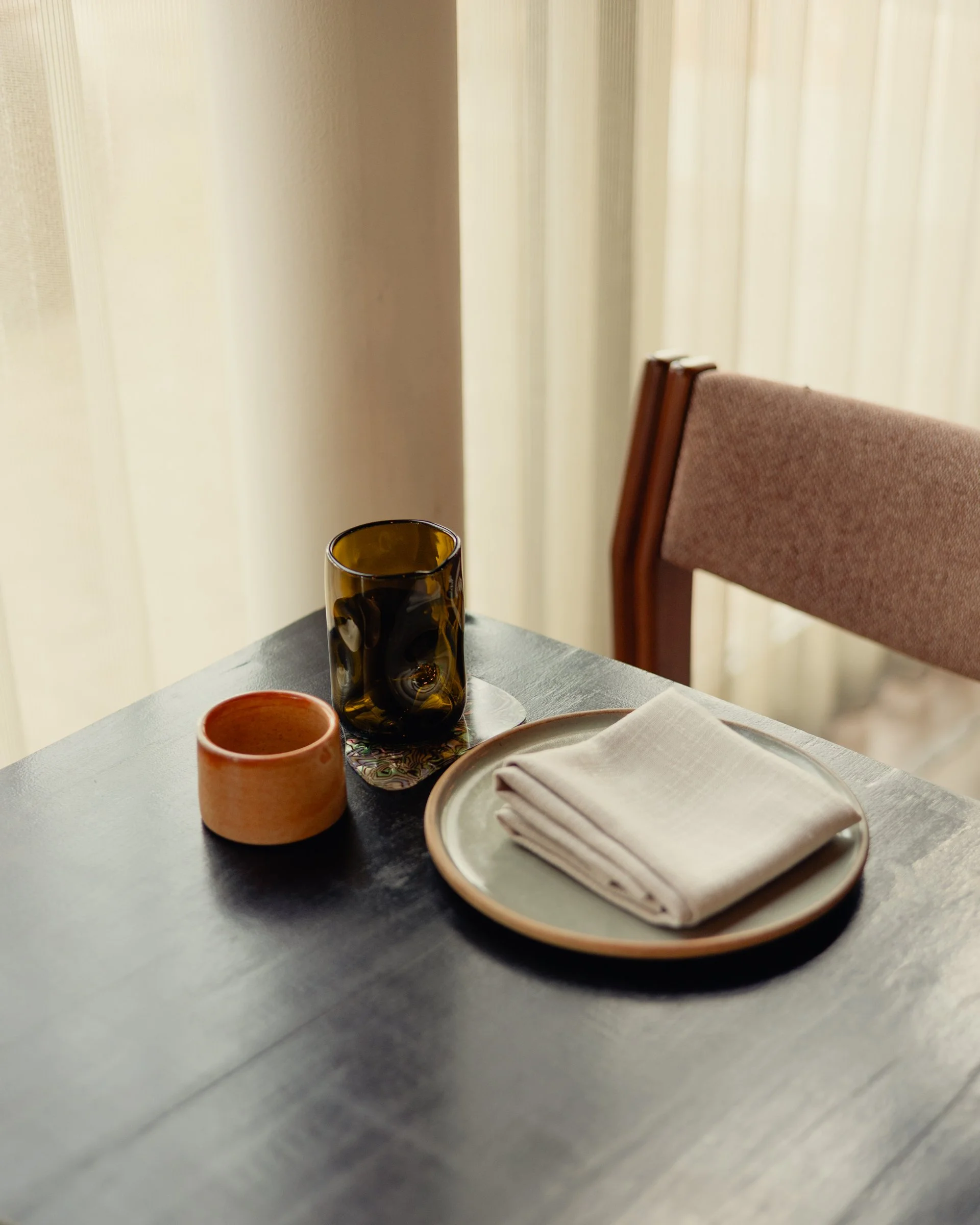 A dark timber table is set with an orange ceramic cup from Chris Harford and water glass by Scott Chaseling. The room is softly lit with cream curtains