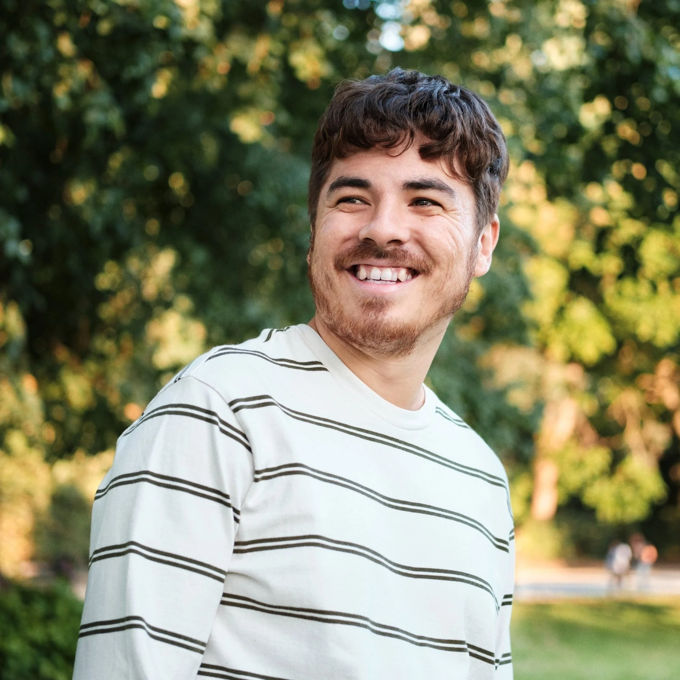 Smiling person wearing a striped shirt outdoors with greenery in the background.