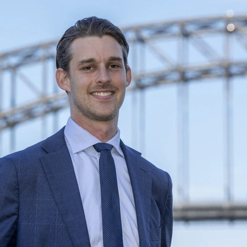 A man in a blue suit and tie smiling outdoors with a steel bridge and blue sky in the background.