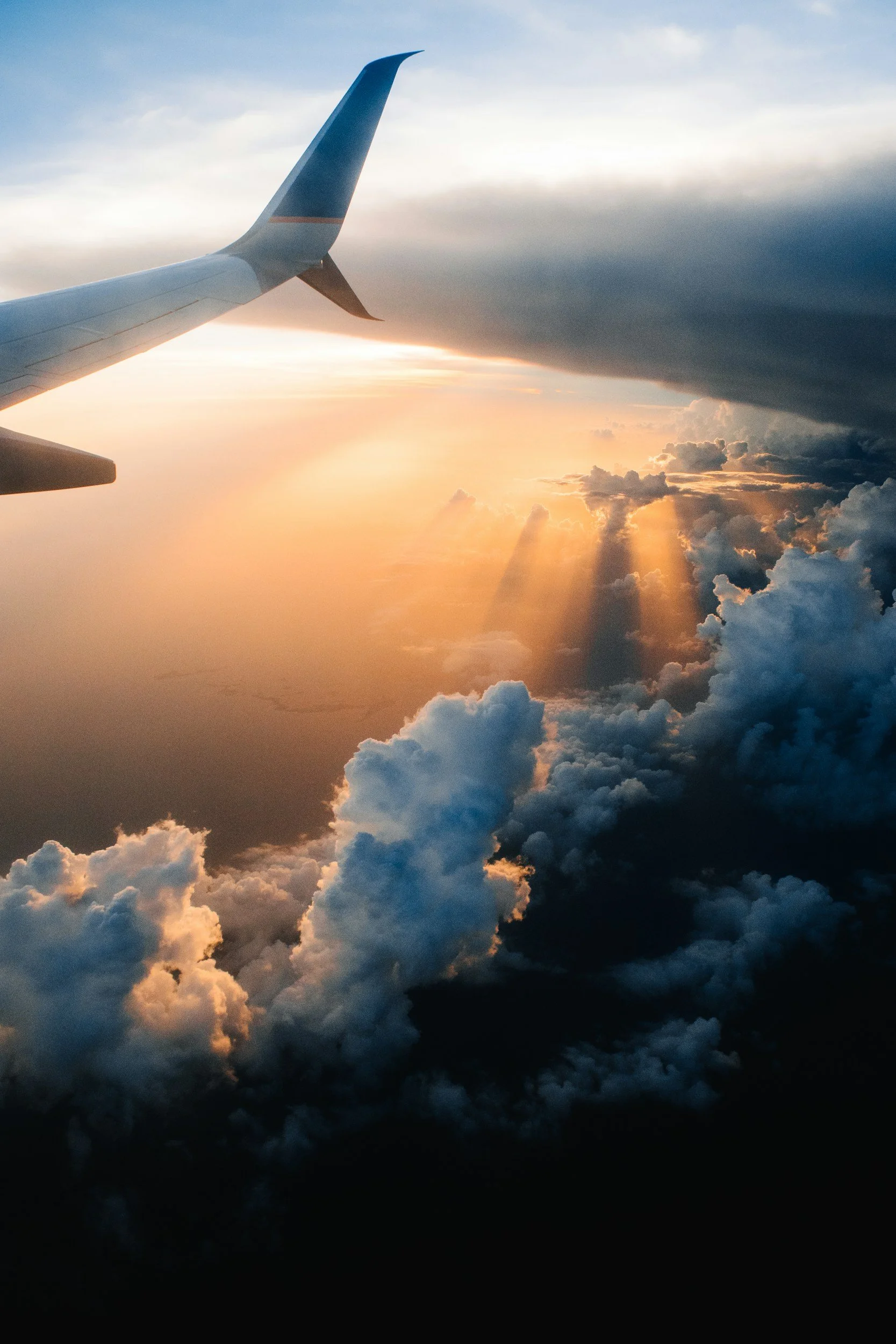 View from an airplane window showing the airplane's wing, sunset sky with rays of sunlight shining through clouds, and a partly cloudy sky with dark and light clouds.