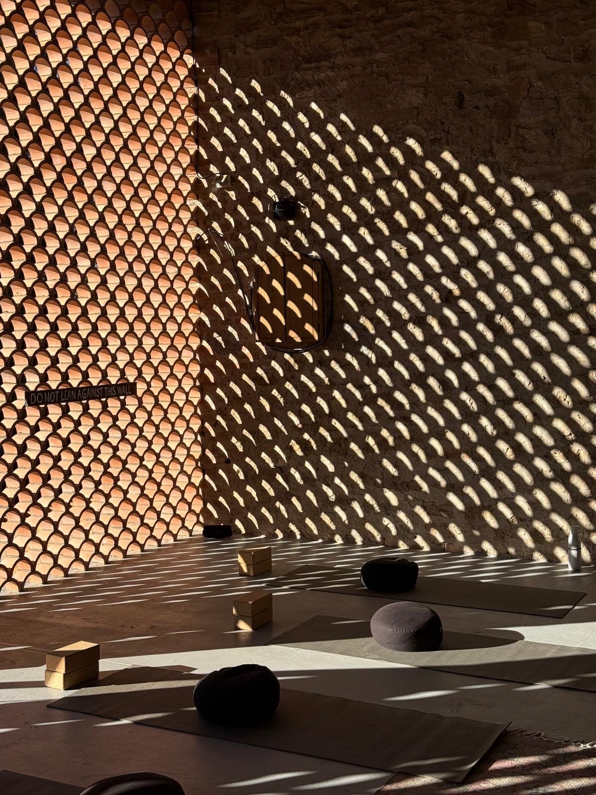 Sunlight streams through a decorative wall, creating a pattern of light and shadow on the wall and floor. The space contains yoga mats, wooden blocks, and rounded cushions, suggesting a yoga or meditation area.