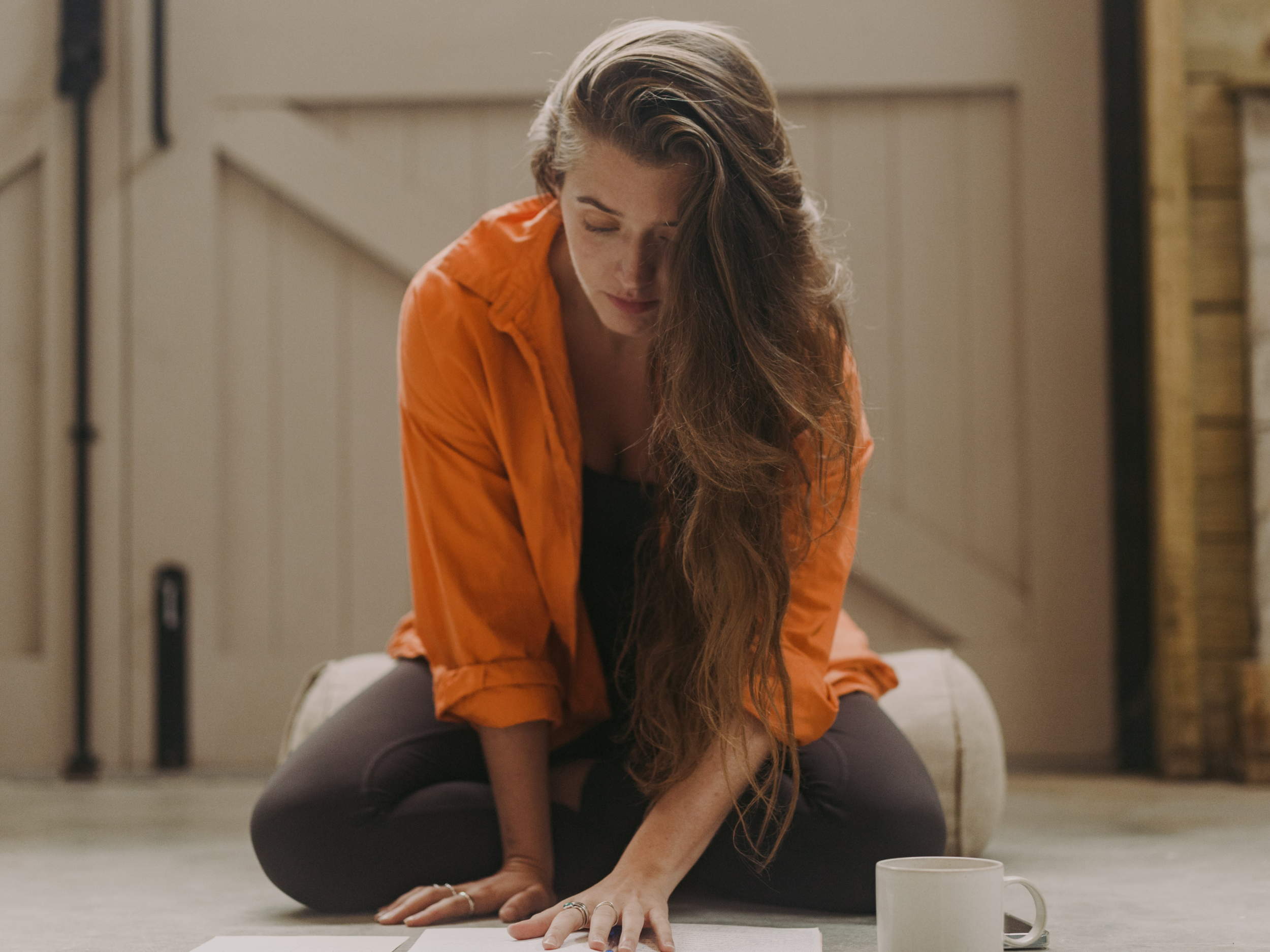 A woman with long wavy brown hair, wearing an orange shirt and gray pants, is sitting on the floor. She is leaning forward and placing her hands flat on a piece of paper or notebook in front of her, with her head tilted downward. There is a white mug on the floor near her right hand. The background shows a neutral-colored wall with some furniture or architectural elements.