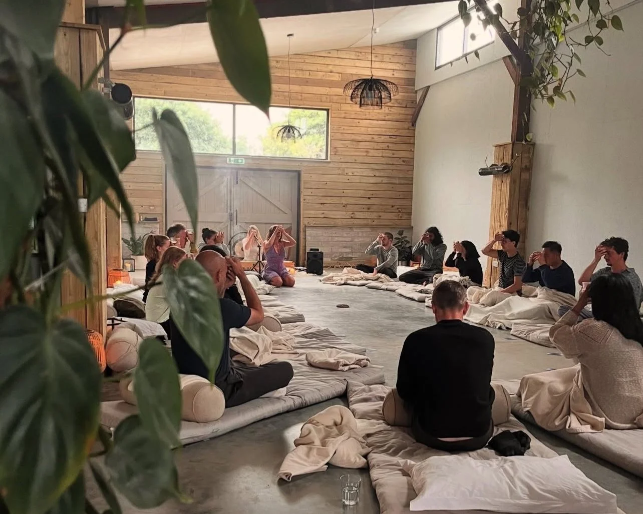 Group of people sitting on mats in a spacious room with wooden walls, participating in a meditation or yoga session.