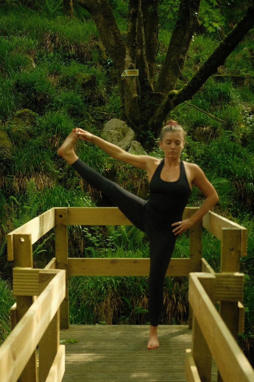 A woman practicing yoga on a wooden bridge in a lush green forest, holding a high leg stretch pose with one arm extended to hold her foot.