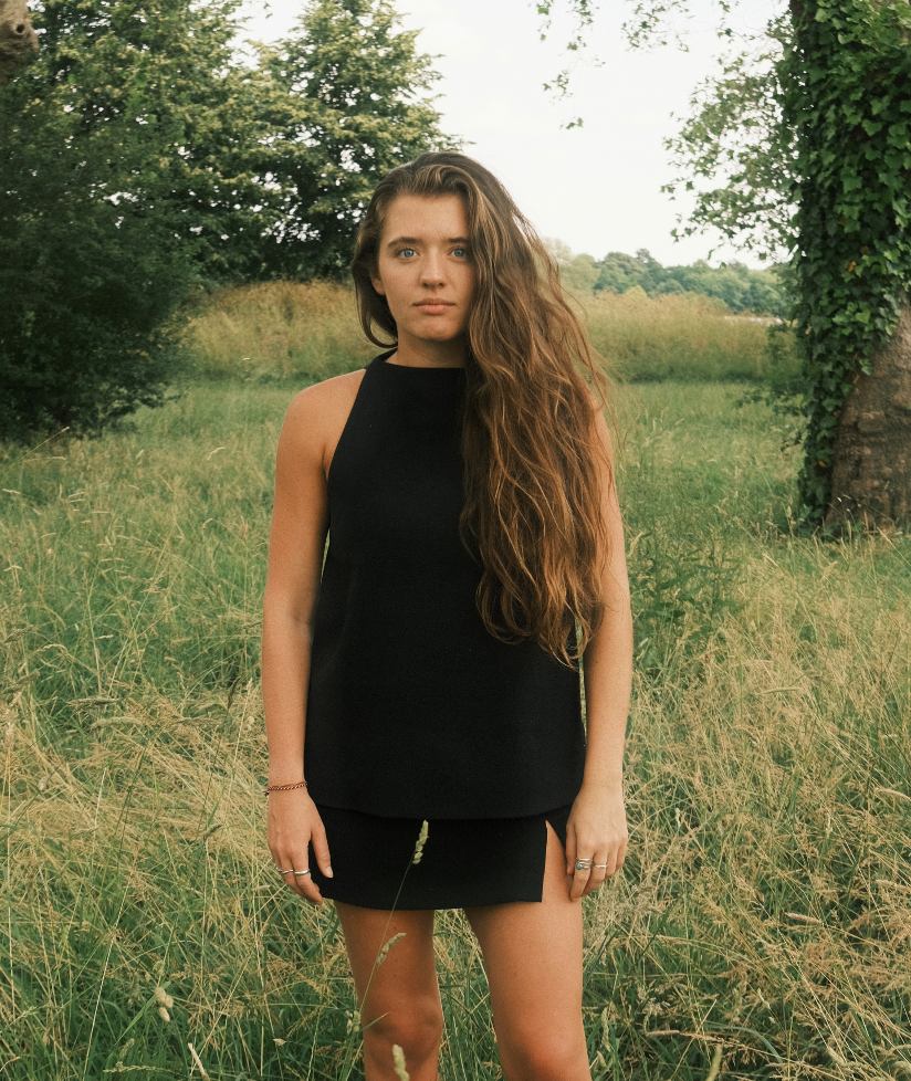 A young woman with long wavy brown hair standing in a grassy field with trees in the background. She is wearing a sleeveless black dress and looking at the camera.