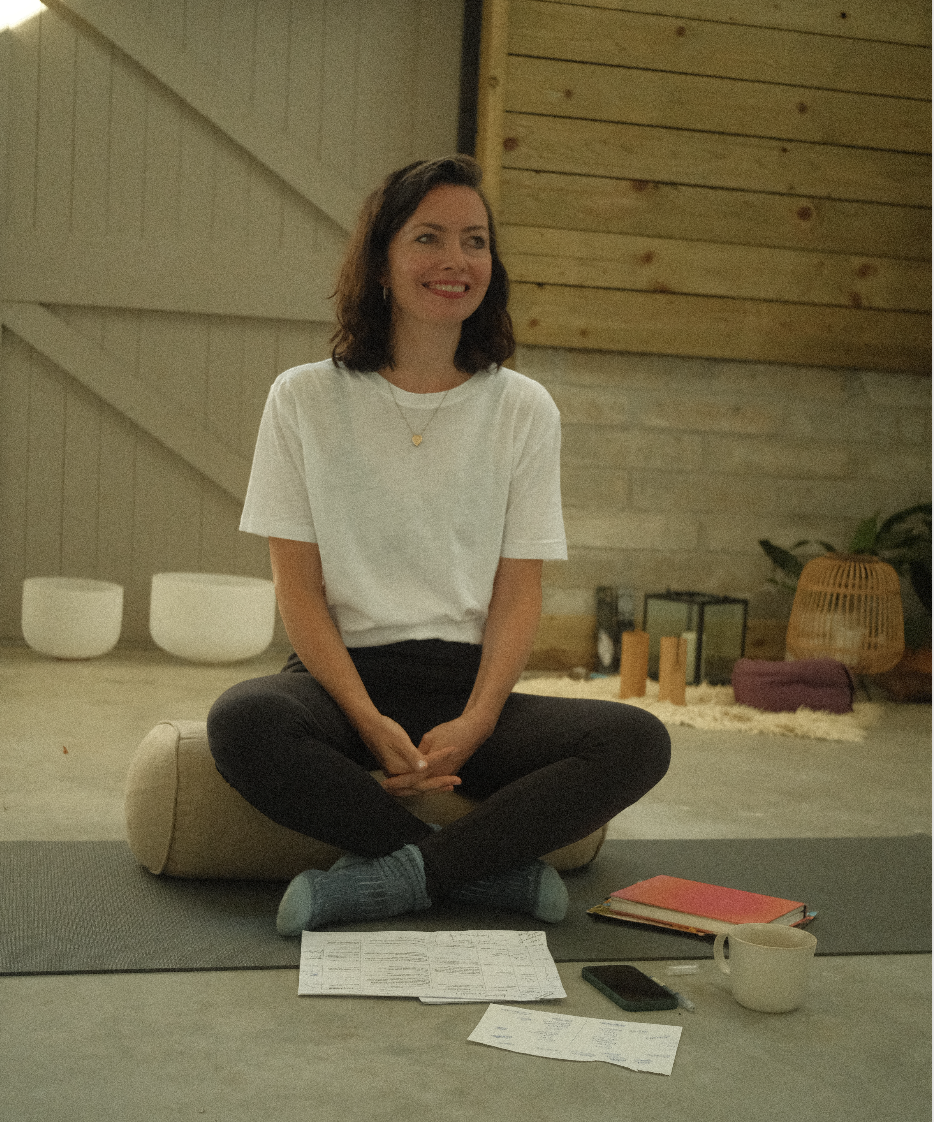 A woman sitting cross-legged on a yoga mat, smiling, in a cozy wooden room with various items including notebooks, a smartphone, a mug, and decorative objects around her.