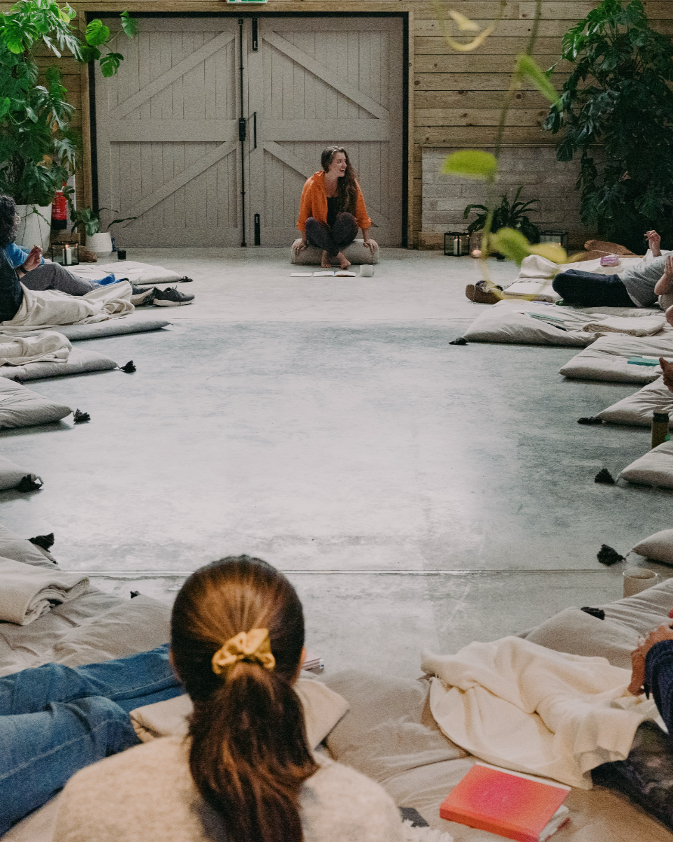A woman with long hair, sitting cross-legged on the floor, leading a group in a meditation or yoga session inside a rustic wooden room with large plants and cushions.