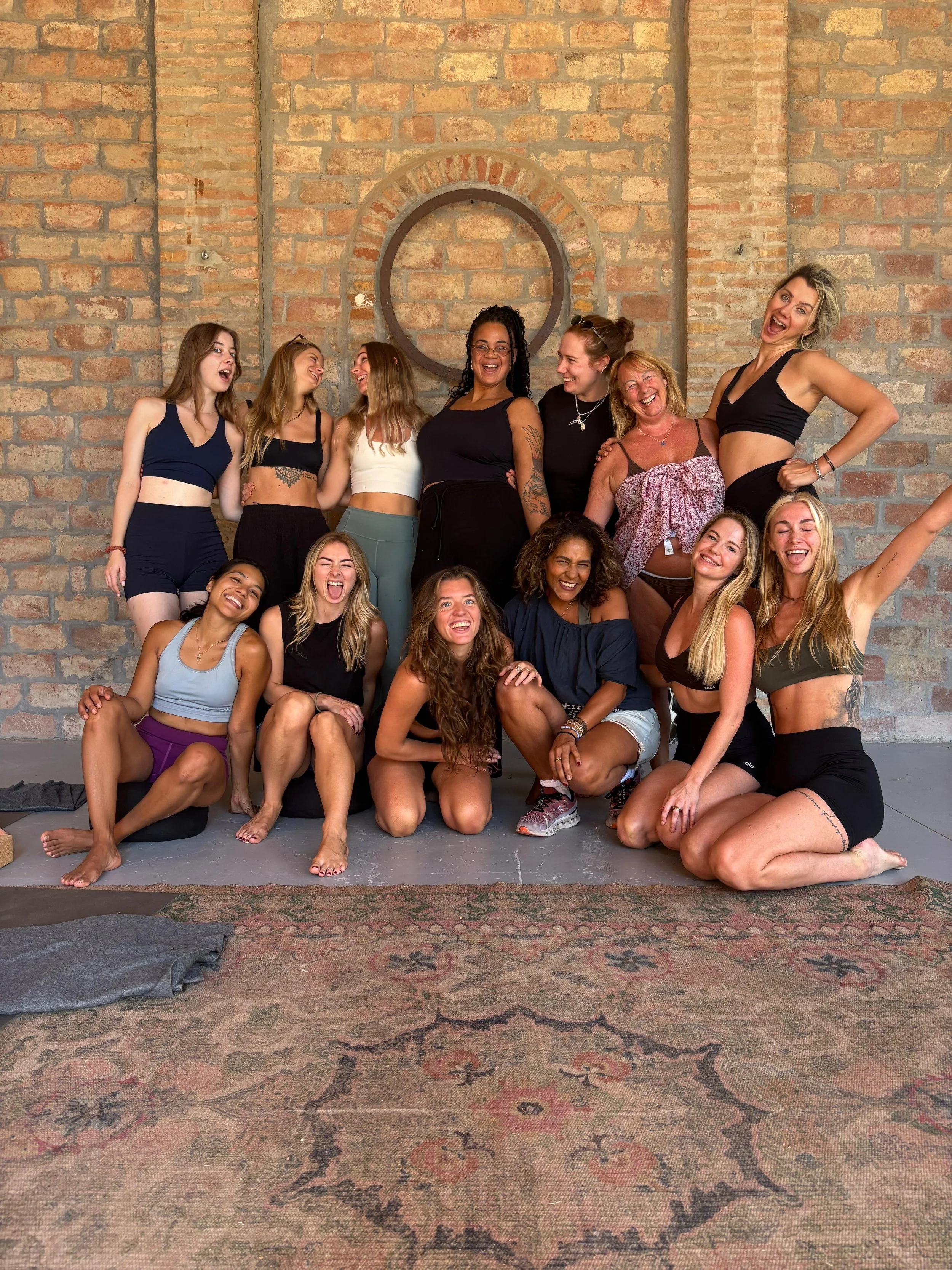 A group of women, smiling and laughing, poses together in front of a brick wall. They are dressed in athletic wear and sitting on a patterned rug, with some standing and others sitting or kneeling, showing a friendly and joyful atmosphere.