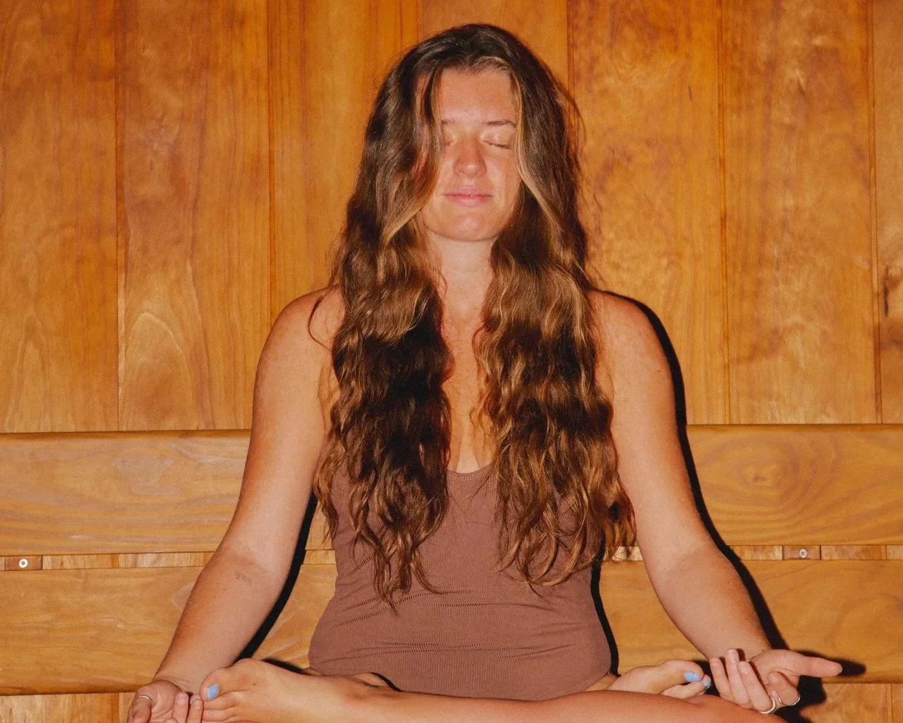 A woman with long, wavy brown hair meditating in a seated yoga pose with her eyes closed, sitting on a wooden bench against a wood-paneled wall.