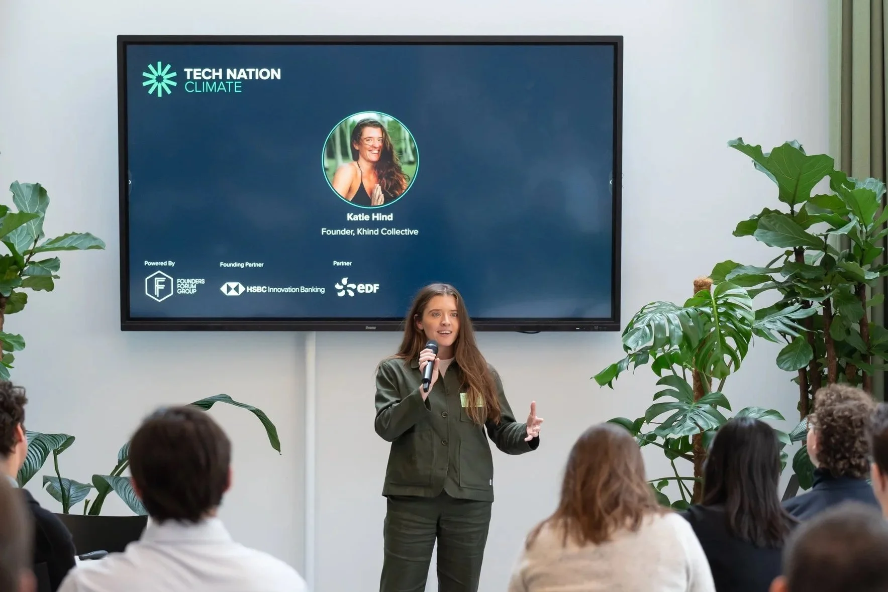 A woman is giving a presentation at a conference with a slide on a large screen behind her. The slide shows a photo of Katie Hind, founder of Khind Collective, under the title 'Tech Nation Climate.' The audience is seated and listening attentively, with large green plants flanking the presentation area.