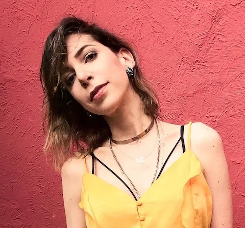 Women with red background and yellow top with her head tilted to one side, half smiling and looking at the camera