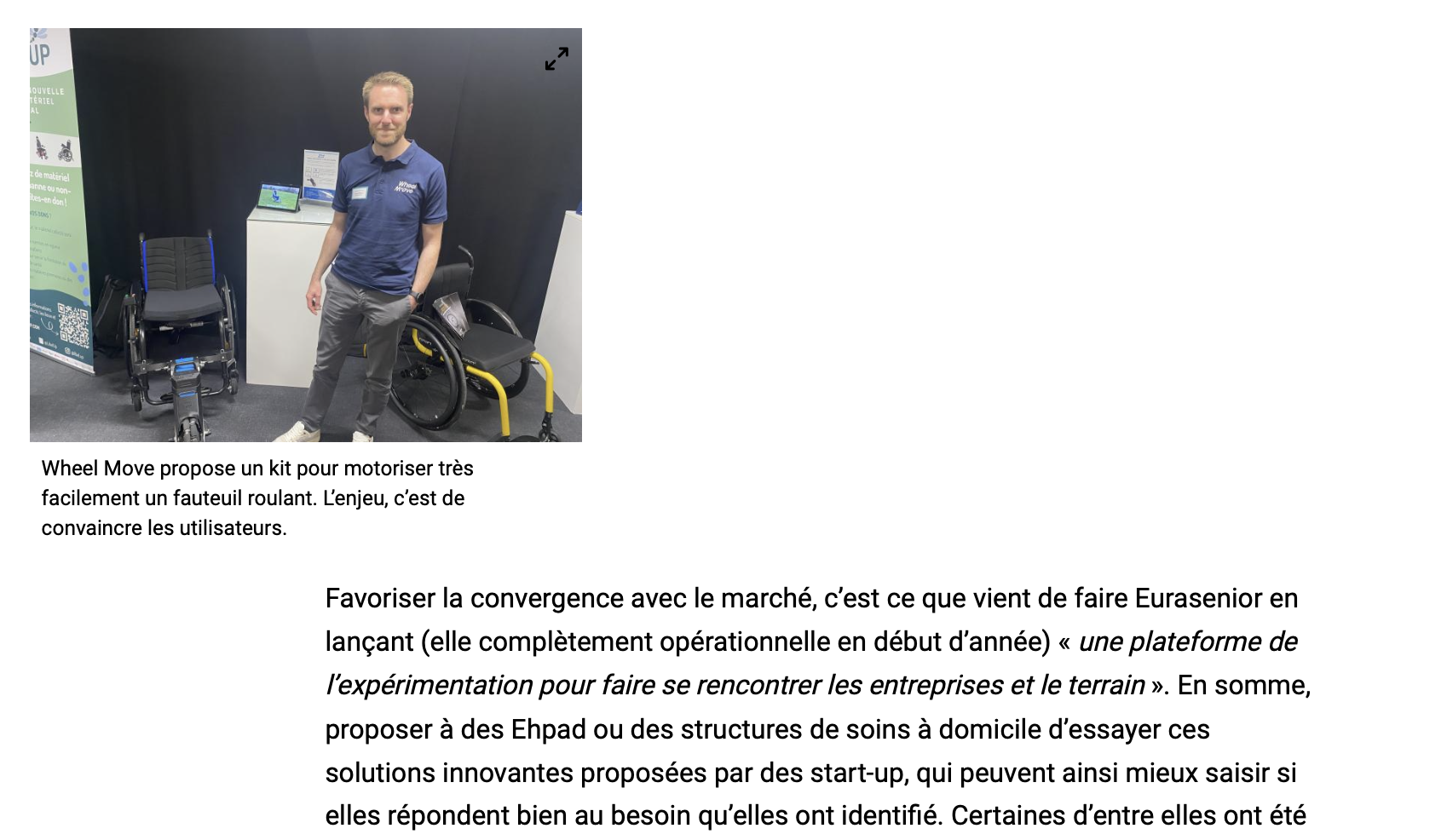 Un homme souriant porte un polo bleue, devant deux fauteuils roulants et un écran dans un espace d'exposition.