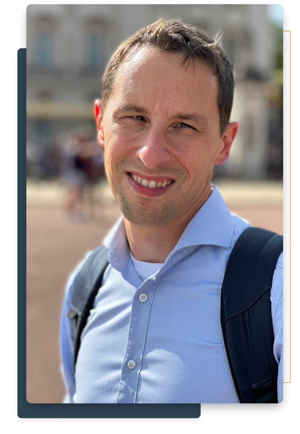 A smiling man with short dark hair wearing a light blue button-up shirt and a backpack, standing outdoors on a sunny day.
